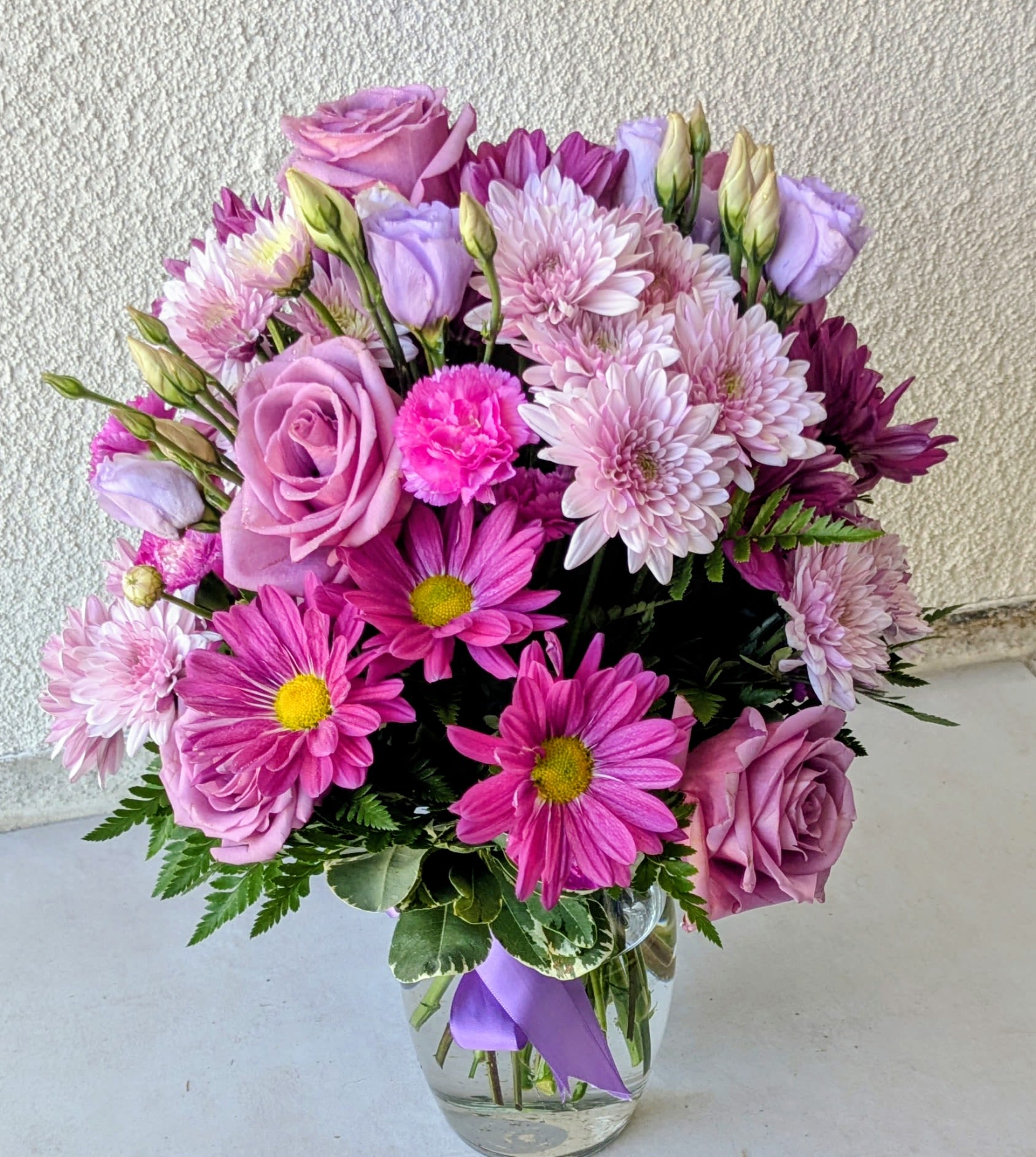 Lavender Garden Delight  - Soft, romantic, and effortlessly elegant, this charming arrangement features a graceful blend of lavender roses, blush pink chrysanthemums, and vibrant magenta daisies. Accented with delicate lisianthus buds and lush greenery, each bloom is thoughtfully arranged to create a full, garden-inspired look. Presented in a clear glass vase and finished with a subtle lavender ribbon, this bouquet is perfect for birthdays, get-well wishes, or simply adding a touch of beauty to any space. Its soothing pastel tones and fresh-picked feel make it a timeless and uplifting gift.