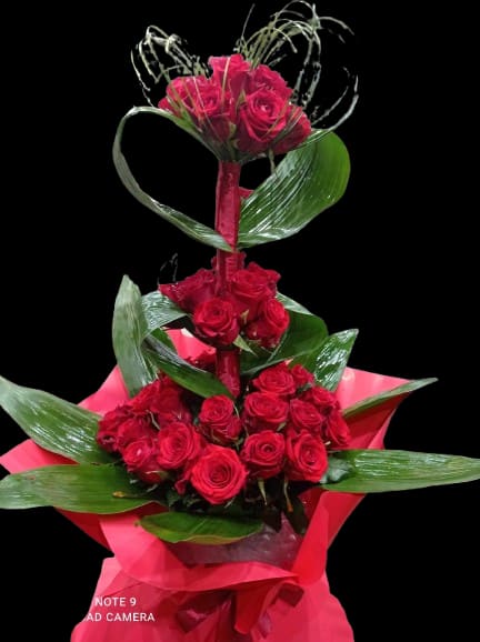 Architect of Love - A dramatic, sculptural rose arrangement that reads as a bold romantic statement - perfect as an anniversary bouquet, Valentine's Day surprise, or a birthday gift for someone you adore. The tall, tiered presentation places tightly clustered red roses in rising groupings so the recipient feels wrapped in blooms; the overall effect is formal and unforgettable, the kind of gift that makes her feel truly seen.  This flower arrangement features deep red roses set against broad glossy leaves and looping decorative foliage that frame the blooms like living architecture. Bright pink decorative wrap at the base provides a vivid contrast to the crimson petals and reinforces the romantic tone, while touches of green foliage add freshness and texture. Sized to make an impression on a mantel, entry table, or bedside, it arrives ready to display - ideal when you want a meaningful, high-impact way to say "I love you."