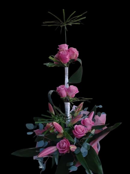 Elevated Grace - Pink roses and pink lily buds rise in a tall, tiered silhouette that reads both dramatic and romantic-an ideal anniversary bouquet or birthday arrangement to make the person you're thinking of feel truly celebrated. The cascading clusters are punctuated with airy green fillers and eucalyptus-like foliage, giving the design graceful movement and a soft, fragrant presence perfect for Mother's Day or any special celebration.  This vertical arrangement features grouped roses and lilies spaced along a single, elegant line; white wrapped stems and decorative ribbon finish the piece for a refined presentation. The color palette of blush pinks, fresh greens, and touches of white creates a timeless, feminine look that reads beautiful in entryways, beside a cake, or on a mantel. Designed for gifting, it arrives ready to display-simply place in a tall vase and refresh water daily to keep blooms looking their best.