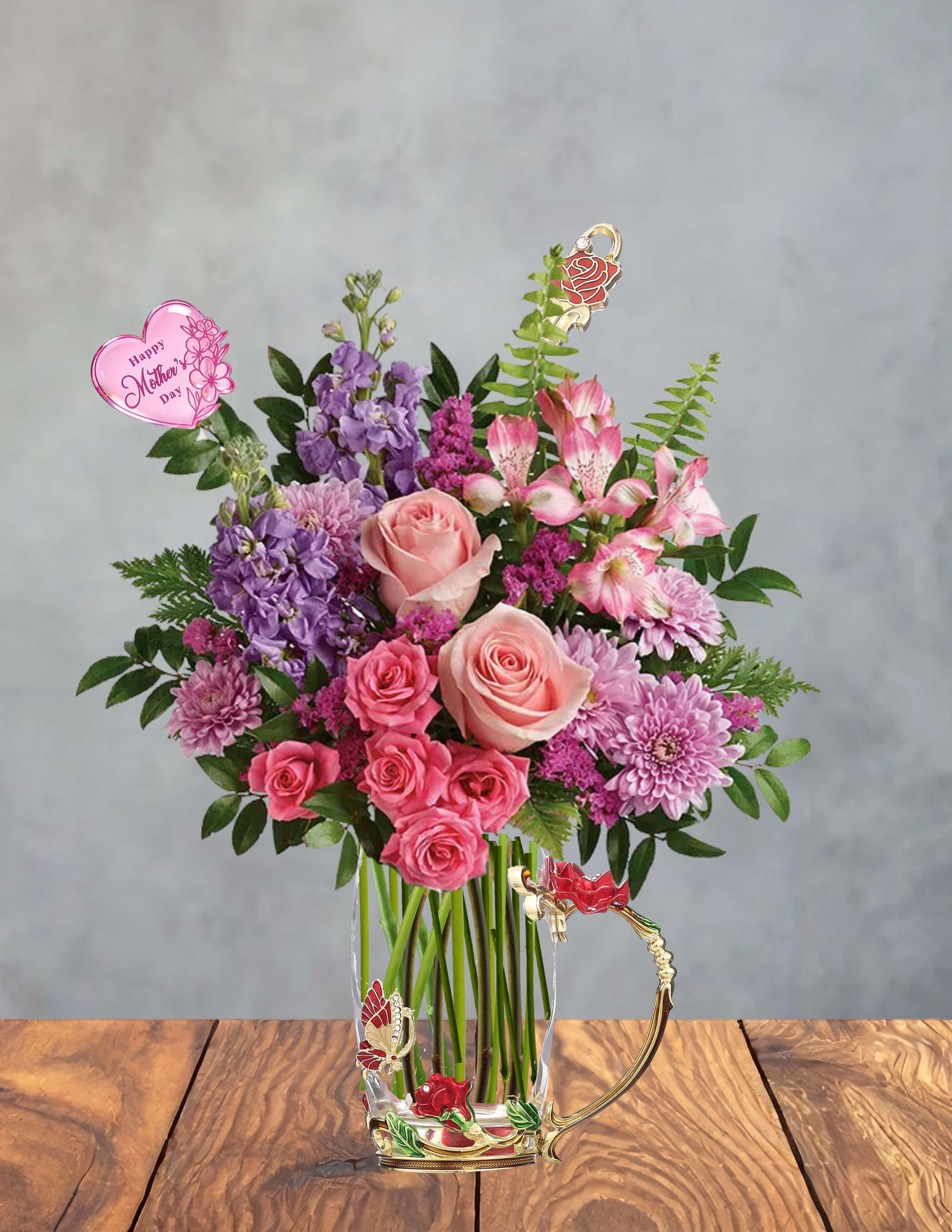 Mother’s Embrace - Soft pink roses and fluttering purple stock set the tone for this Mother's Day bouquet and gift set, a birthday or thank-you arrangement designed to make the person you're thinking of feel truly cherished. The upright, full design combines lavender chrysanthemums and pink alstroemeria with visible stems in a clear glass vase for a lush, layered look; small heart-shaped picks - including one that reads "Happy Mother's Day" - add a sentimental, celebratory touch that says "you matter" without words.  This arrangement pairs with a decorative hot-or-cold glass cup and matching spoon, both set in a gold-toned metal frame and finished with a red rose motif, butterfly accents, and tiny green enamel details for a romantic, gift-box feel. The mix of roses, alstroemeria, chrysanthemums, and stock delivers soft pinks, purples, whites and a touch of green for a feminine palette that fits birthdays, Mother's Day, anniversaries, or an everyday "thank you." The glass vase and metal-accented cup make the bouquet ready to display; simple care-fresh water and a cool spot away from direct sun-keeps blooms looking their best so the recipient enjoys the moment longer.