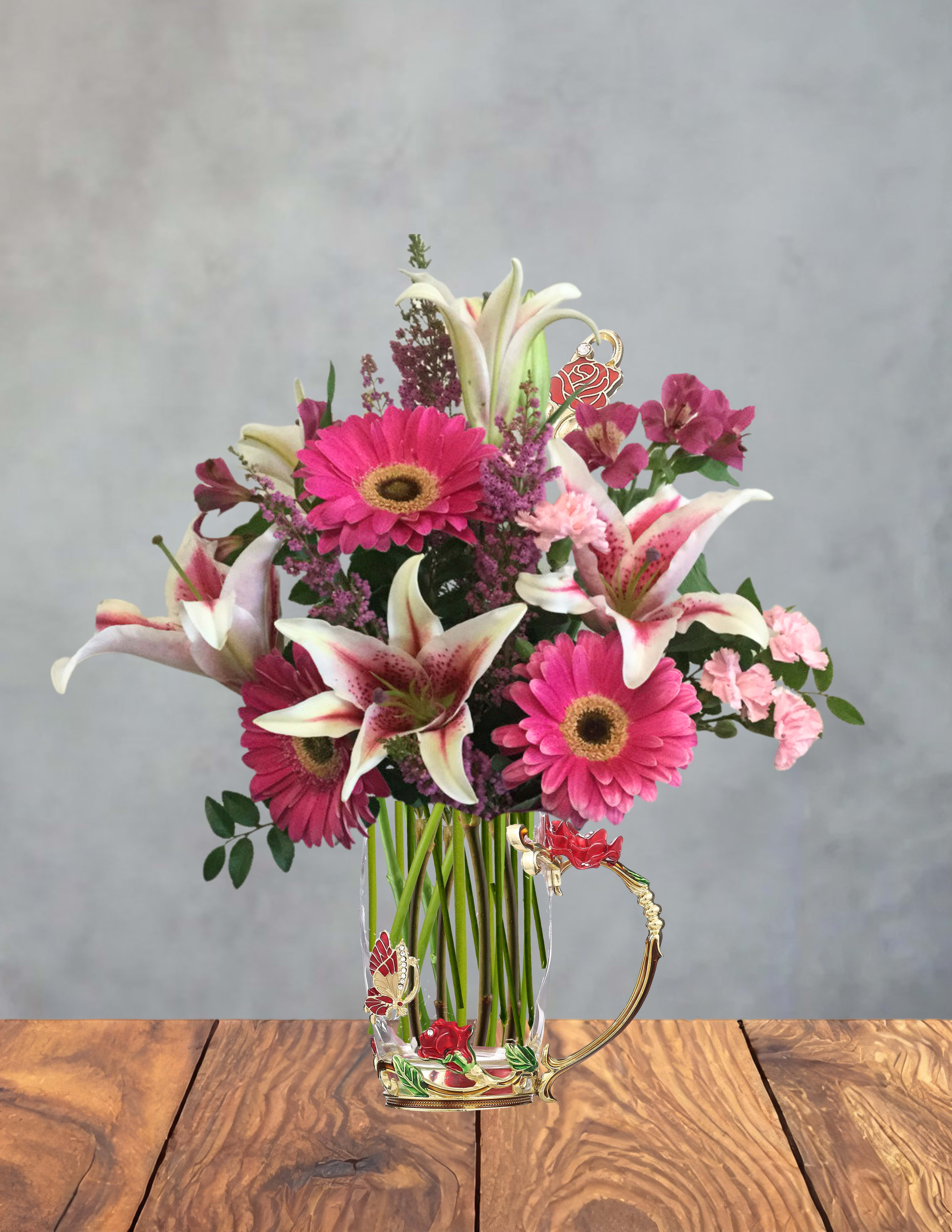 Heart of her garden - Bright pink gerbera daisies and white-and-pink lilies set the tone in this birthday bouquet-style flower arrangement, the perfect spring celebration or thank-you gift that feels both cheerful and slightly romantic. Designed as a gift for the person you're thinking of, the mix of vivid pinks, rosy accents and purple filler flowers creates an immediate visual lift-she'll feel seen and celebrated the moment she opens it. This arrangement also reads as a lovely anniversary or "just because" surprise for someone who loves feminine, decorative touches.  The flowers are gathered in a clear glass mug finished with an ornate gold-toned floral handle and metal embellishments, paired with a matching decorative spoon and butterfly accents for a keepsake touch. You'll find gerbera daisies, lilies, small pink alstroemeria, roses and purple filler blooms arranged with greenery for contrast; the glossy, ornamental cup features red rose motifs and tiny rhinestone-like highlights that make the presentation feel special. Sized as a gift-ready tabletop piece, it arrives assembled in the decorative glass cup so no vase is needed-light trimming of the stems and fresh water will keep blooms looking their best for several days.