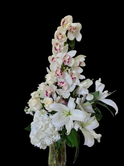 Orchid Grace - Cascading white dendrobium orchids rise above large white lilies in this tall flower arrangement, creating a graceful, vertical silhouette perfect for a sympathy tribute, wedding display, or a refined celebration bouquet. The arrangement reads as an elegant everyday centerpiece that still feels ceremonial-ideal for the person you want to honor or comfort with a calm, uplifting presence.  A rounded cluster of white hydrangea anchors the base while soft pink accents in the orchid centers and pale roses add a hint of warmth against the predominantly white and green palette. Arranged in a clear glass vase, the layered composition emphasizes height and airiness; the mix of textured hydrangea, sculptural orchids, and fragrant lilies gives visual depth and a gentle scent. This is the kind of arrangement that makes someone feel seen and soothed-order as a thoughtful sympathy expression, wedding accent, or meaningful everyday gift.