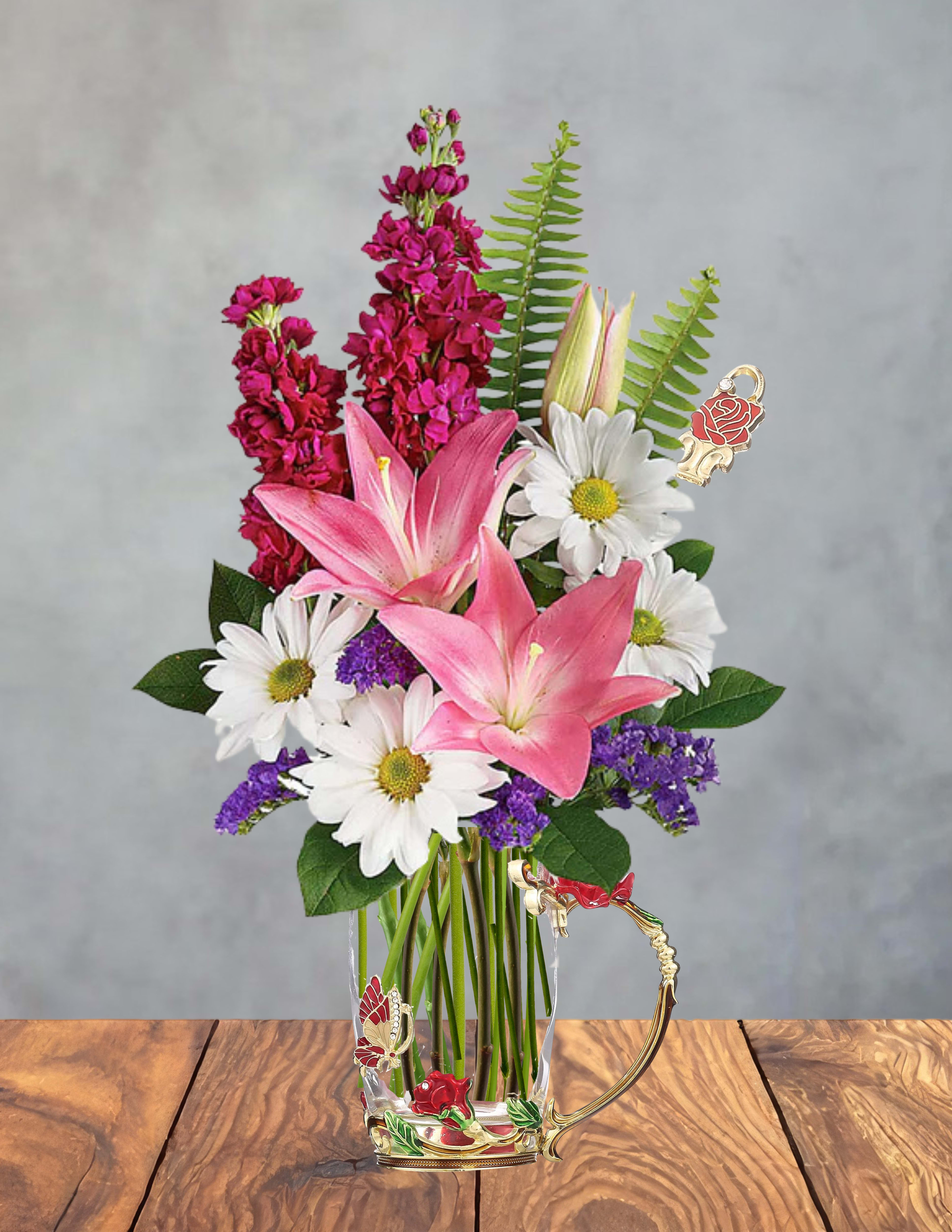 Mom’s Garden Glow - Two large pink lilies take center stage in this Mom's Garden Glow flower arrangement, giving it a romantic, garden-fresh presence perfect for Mother's Day, a birthday, or a heartfelt thank-you bouquet. White daisies and clusters of small purple filler flowers soften the composition while tall magenta spikes rise behind the main blooms to create a layered, upright silhouette that catches the eye from every angle - the kind of gift that makes the person you're thinking of feel truly celebrated.  Arranged in a clear glass vase with a decorative handle and ornate floral accents, the presentation reads as both elegant and ready-to-gift. Colors include pink, white, purple, and green for a bright, cheerful palette; textures range from the lilies' silky petals to the daisy centers and delicate filler clusters, giving the bouquet a lively, garden-like feel. Ideal for spring occasions or everyday gifting, this arrangement arrives assembled and vase-ready - care instructions: keep water fresh, trim stems every few days, and place away from direct heat to extend bloom life.