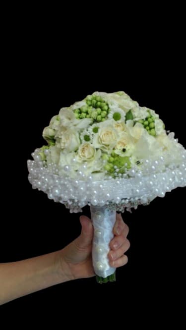 Pearl Bloom - A compact, rounded hand-tied bouquet that reads bridal and timeless-white and cream roses mingle with daisy-like blooms and soft clustered filler for a formal wedding bouquet or ceremony arrangement. The predominantly white palette with small green accents creates a fresh, elegant look that makes the person you're thinking of feel cherished and gracefully celebrated.  Cream-white roses provide a classic focal point while the mixed small daisies and textured filler soften the silhouette; the stems are finished with a wrapped handle and pearl trim for a distinctly bridal presentation. Sized to be carried comfortably, this bouquet suits brides, attendants, or anyone marking an important ceremonial moment; keep stems in fresh water until presentation and handle the wrapped base gently to preserve the pearl detailing.
