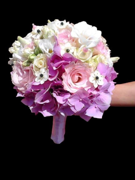 Sugar Bloom - A tightly rounded handheld bouquet in soft pinks, whites, and lavender makes a quietly romantic statement-perfect as an anniversary bouquet, a spring celebration gift, or a bridal-ready posy. The compact, formal shape reads as polished and feminine, the kind of arrangement that will make the person you're thinking of feel cherished and truly seen.  Large rose heads set the tone, layered with elegant calla lilies and smaller white accent blooms that add delicate, airy texture and contrast. The color palette of blush pink, pure white, and gentle purple keeps the look cohesive and refined. Finished with a wrapped bouquet handle and a neat ribbon, this arrangement arrives ready to carry or present without fuss.  Give this bouquet when you want to mark a meaningful moment-a vow renewal, a birthday, or simply to celebrate someone special this spring. Hand-delivered and designed to sit comfortably in the recipient's hands, it's easy to order and makes an immediate, flattering impression; keep flowers in fresh water and out of direct sun to extend their beauty.