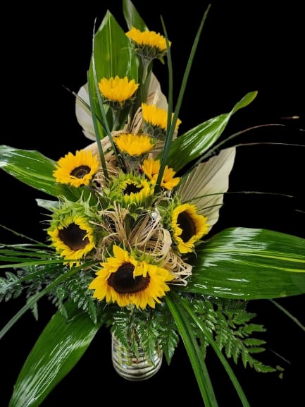 Summer Day - Bright, cheerful sunflowers take center stage in this yellow, green, and white flower arrangement - a sunny birthday bouquet or congratulations gift that immediately lifts the room. Clustered at varying heights in a clear glass vase, the golden heads read as joyful and confident; the arrangement is perfect for celebrating milestones or simply letting someone know you're thinking of them.  Large glossy tropical leaves and fern-like greenery give the bouquet a bold, layered structure, while pale raffia woven near the center adds a rustic texture that softens the composition. The clear glass vase showcases the stems and keeps the look fresh and modern; this everyday arrangement suits desks, kitchen tables, or entryway consoles. Recipients will feel genuinely cheered by the vibrant color contrast and lush foliage - order in standard sizing and include simple care: keep water fresh, recut stems every few days, and display out of direct heat for longest enjoyment.