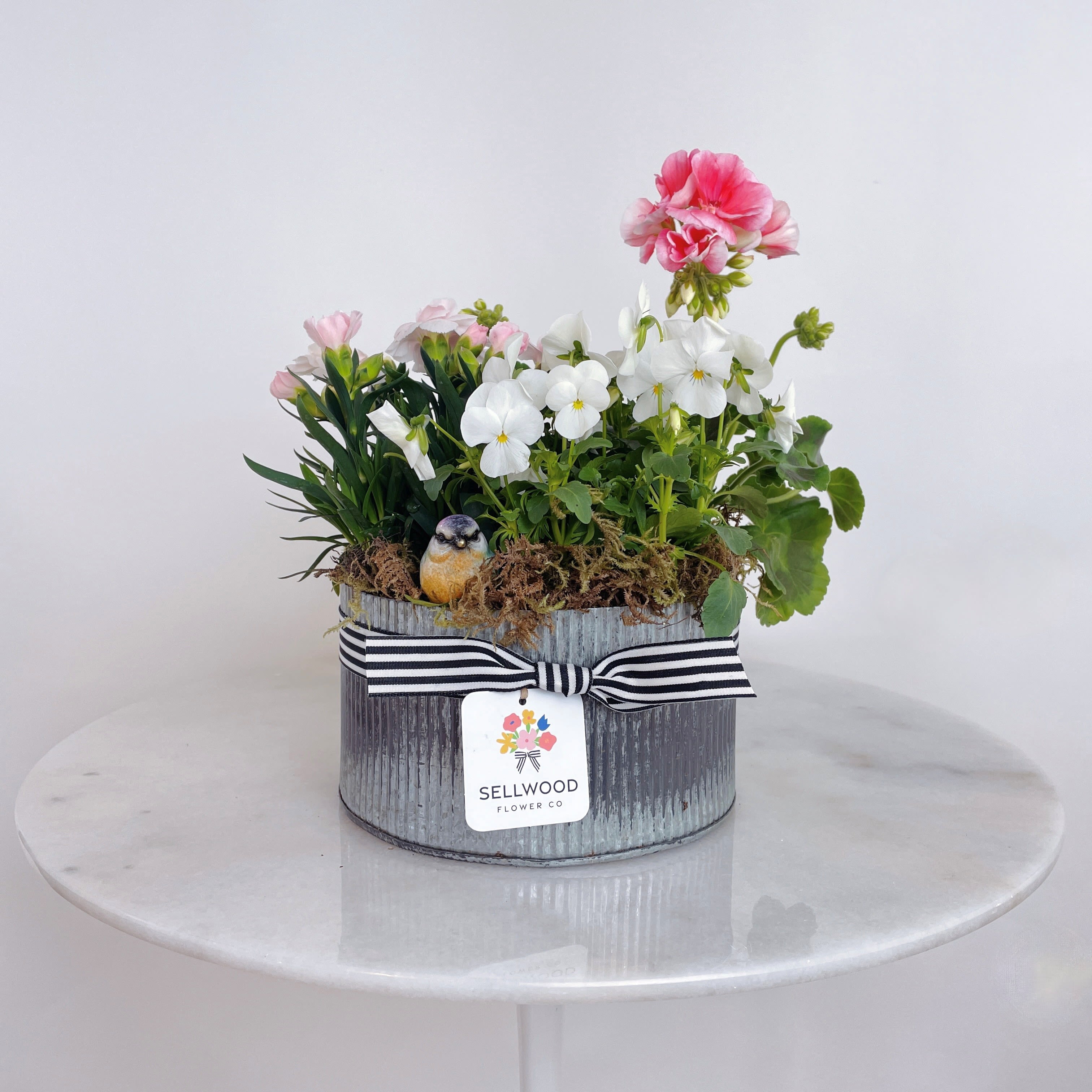 Oval galvanized planter with pink and white flowering plants, moss, and a small bird figurine with striped ribbon.