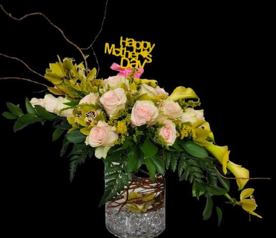 Dear Mom  - Pale pink roses, sunny yellow calla lilies, and delicate orchid blooms come together in a low event arrangement made to make Mom feel celebrated - perfect as a Mother's Day centerpiece, a bridal shower focal piece, or a springtime gift for the woman who deserves something special. The combination of soft pinks, bright yellows, clean whites, and lush greens creates a fresh, uplifting palette that reads as both elegant and joyful for anniversaries or family gatherings.  The flowers are arranged low and full in a clear glass vase, layered over abundant dark foliage that gives the composition depth and a slightly dramatic contrast. Decorative branching twigs weave through the design for texture and height without overpowering the compact silhouette, while a festive yellow "Happy Mother's Day" topper adds a cheerful, personal touch. Sized to sit comfortably on a dining table or reception desk, this arrangement balances visual impact with easy placement.  Designed for the person you're honoring, this piece sends a message of appreciation and warmth-she'll feel truly seen when this arrives. For best longevity, keep the vase water fresh and away from direct heat; if ordering for an event, allow time for delivery and placement so the arrangement can be enjoyed at its peak.
