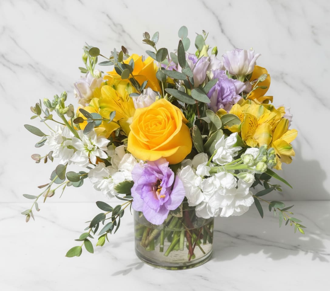 Low glass vase of yellow roses, lavender lisianthus, white stock, and yellow alstroemeria