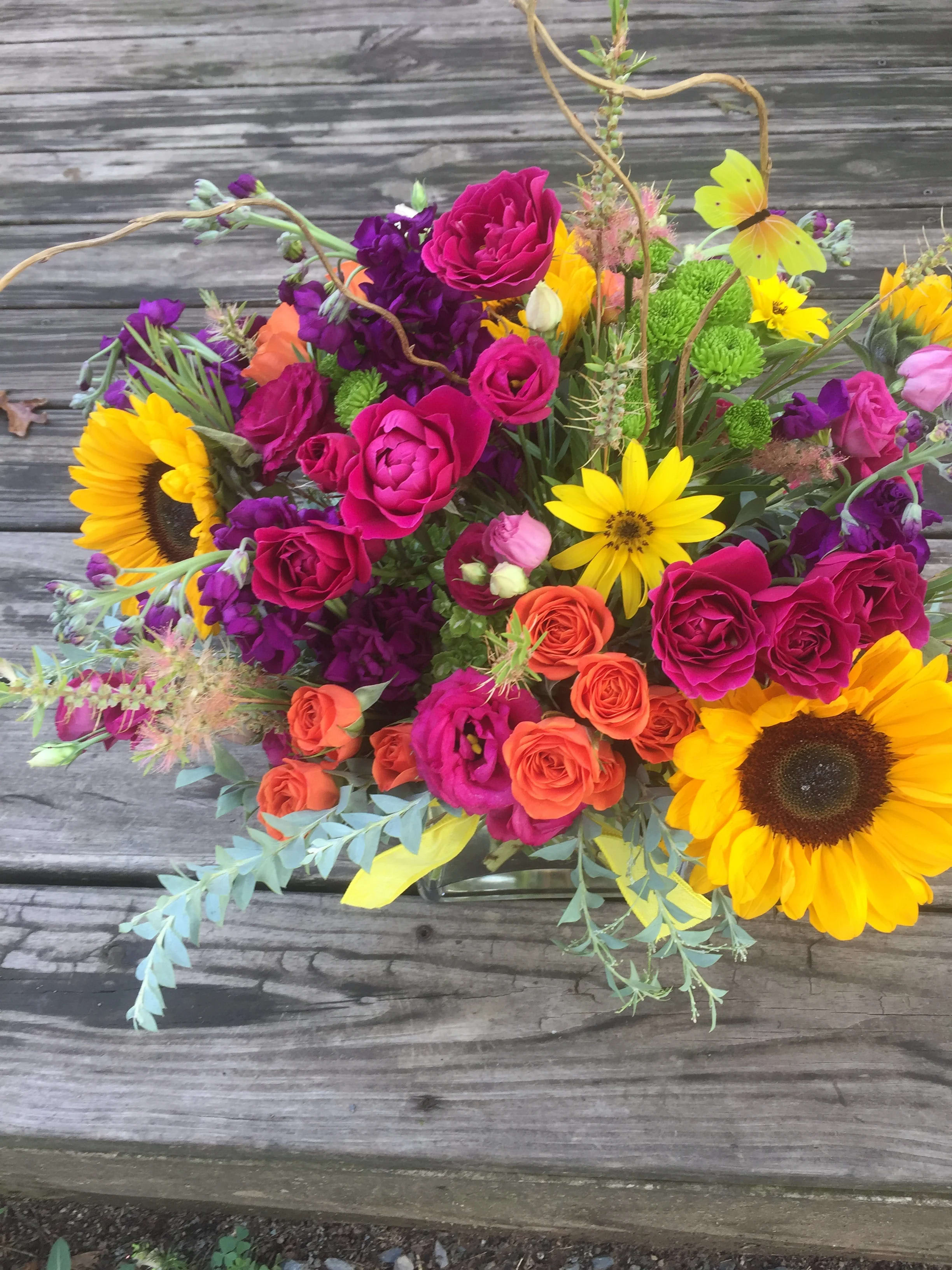Bright mixed arrangement with sunflowers, hot pink and orange roses in a clear glass vase on a wooden surface