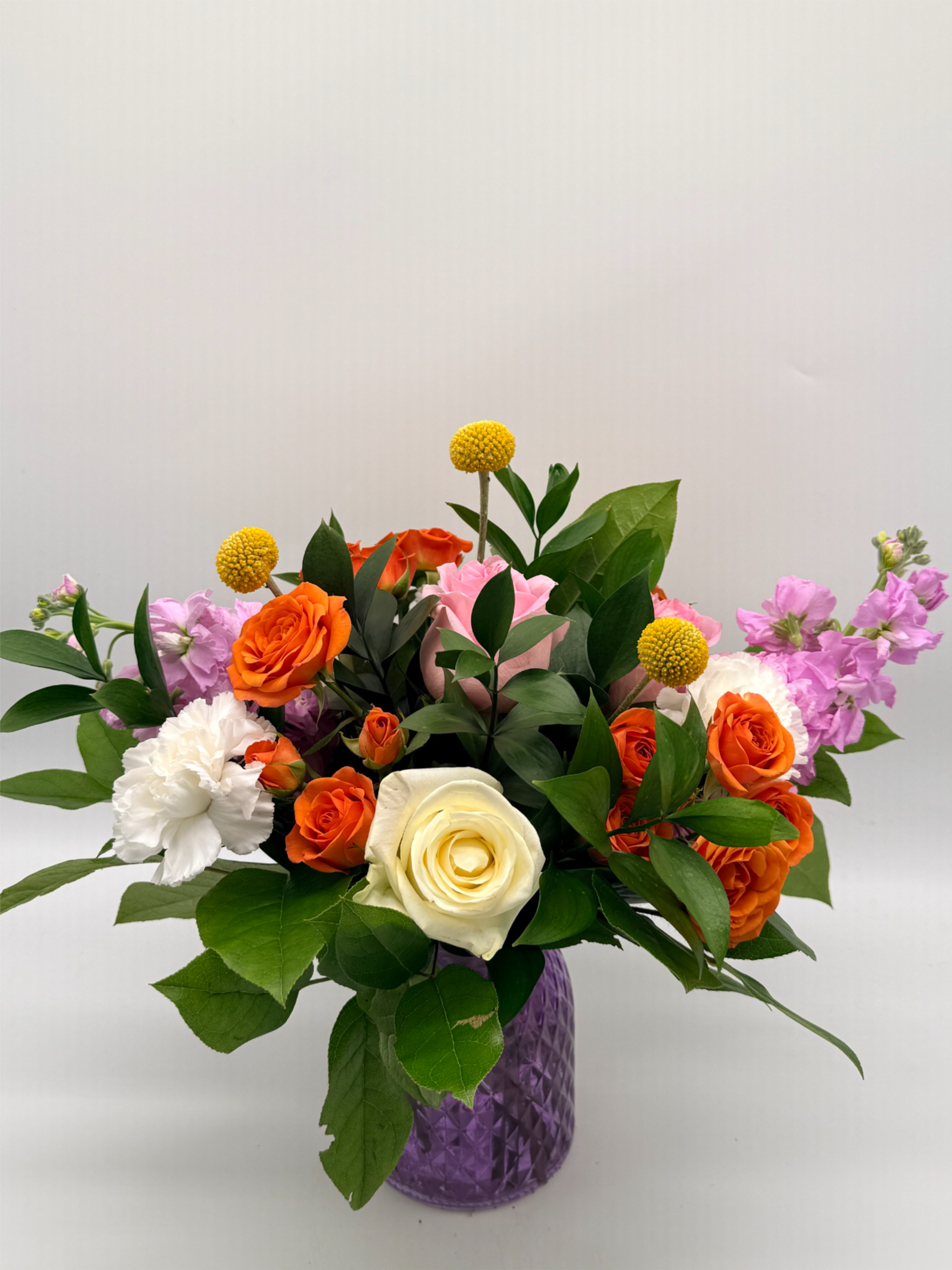 Colorful arrangement of orange roses, white rose and carnation, and pink blooms in a purple glass vase