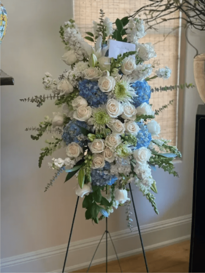 Forever Mine - A tall, formal standing spray arranged on a black metal easel, this sympathy flower arrangement opens with serene white roses and lilies set against clusters of blue hydrangeas and pale green chrysanthemum-like blooms. The cool white-and-blue palette and refined vertical silhouette create a calm, commemorative presence perfect for sympathy, memorial services, or other formal indoor occasions-the kind of arrangement that helps the person you're honoring feel remembered with dignity.  The composition is dense and layered, combining rounded hydrangea clusters and roses with the trumpet shapes of lilies and airy branching foliage for balance and movement. Trailing greenery softens the edges while the black metal easel gives it an elevated, ceremonial look; a small card is tucked near the top for your message. Designed to read well from a distance, the spray's full height and formal profile make it suitable for funeral chapels, memorial gatherings, or as a statement tribute at home.