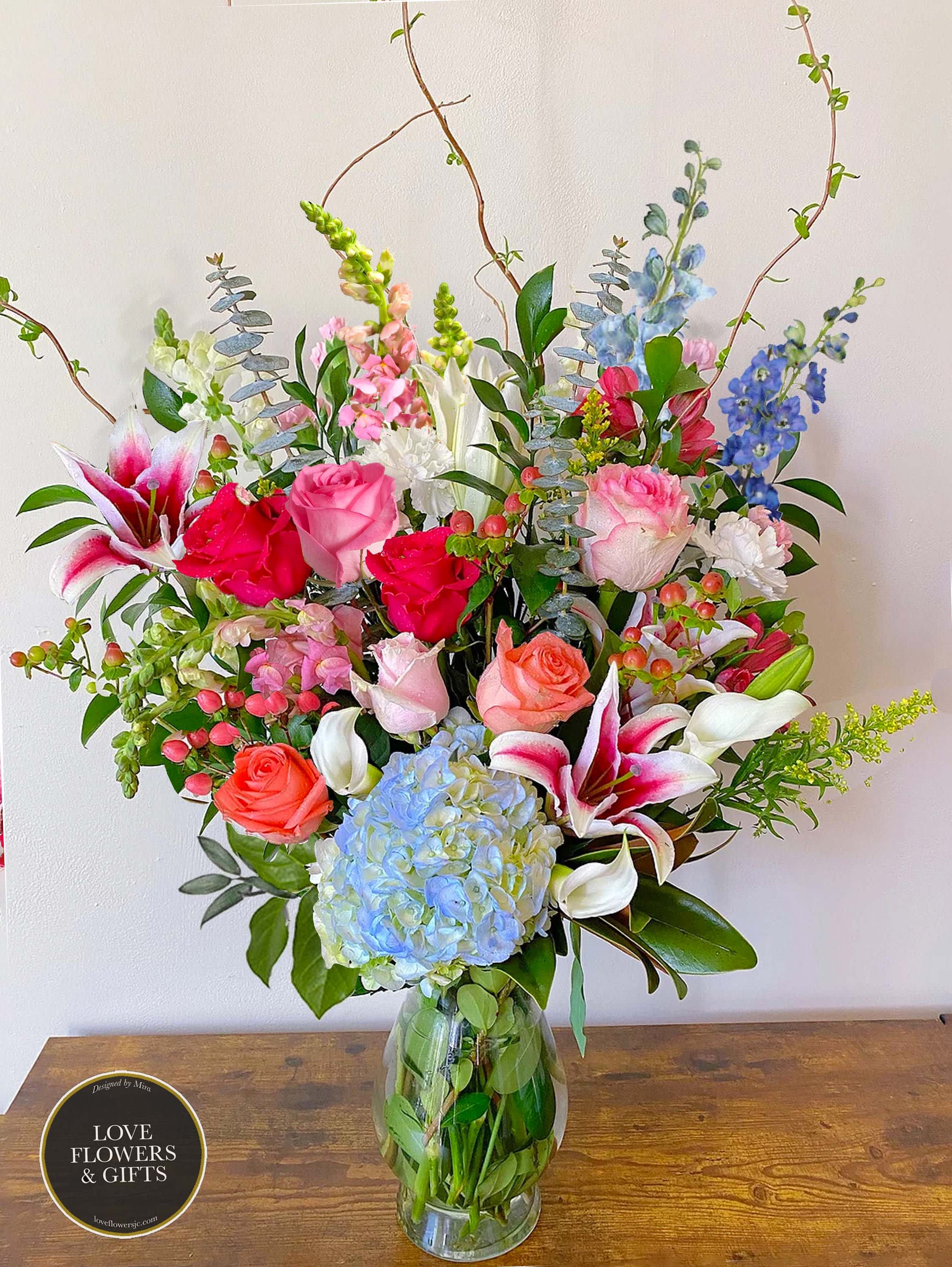 Tall mixed arrangement of pink roses, lilies, blue hydrangea and assorted blooms in a clear glass vase