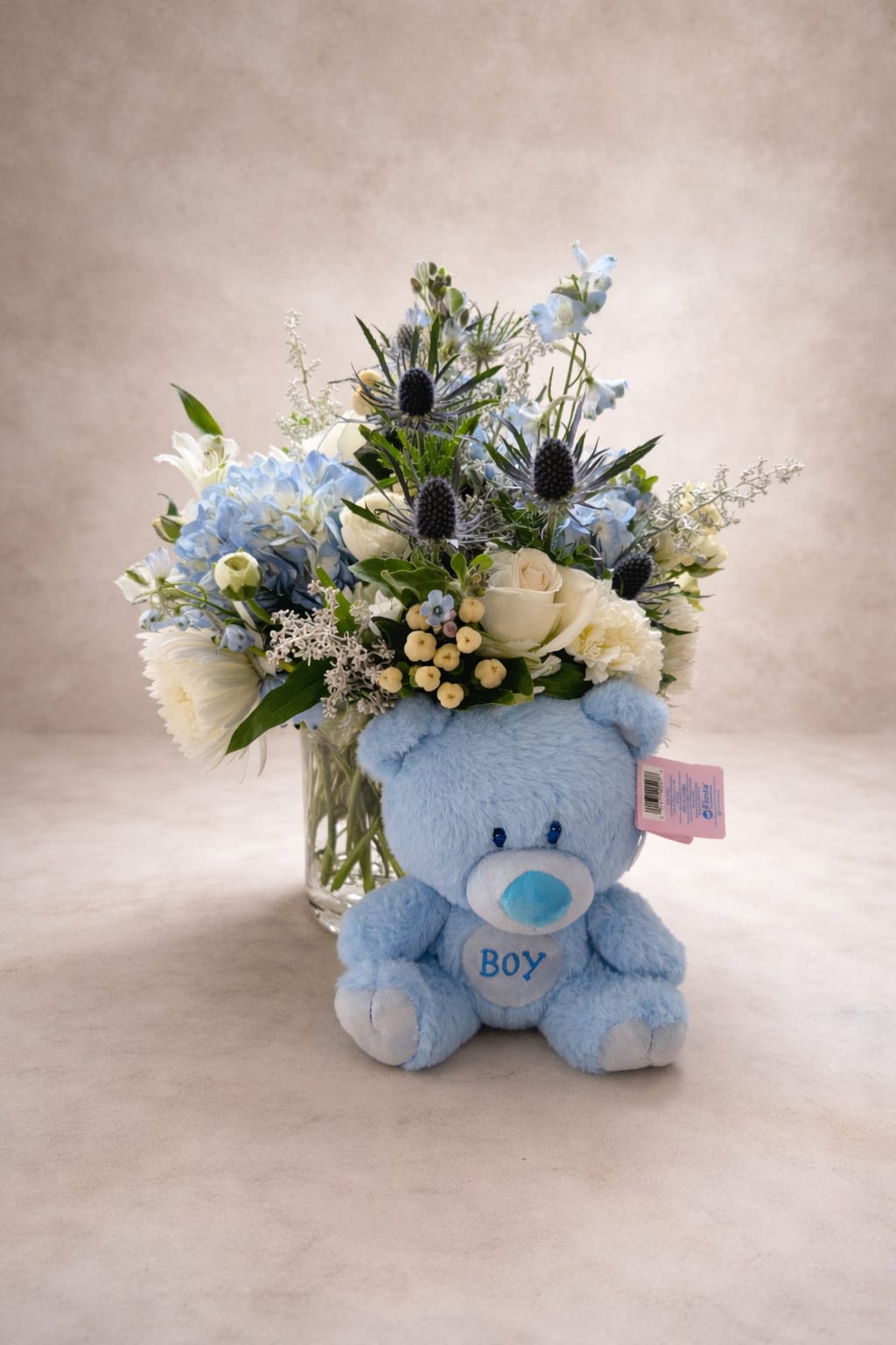 Blue baby boy - Pale blue hydrangeas and soft white roses create a gentle, celebratory look in this baby boy flower arrangement-perfect as a newborn gift or baby shower centerpiece. The arrangement's baby-blue emphasis and airy palette send a clear message of welcome and tenderness, so the new parent will feel truly seen and celebrated.  Creamy white carnations, light blue accent blooms and delicate dendrobium orchids fill a rounded, full composition, while darker spiky, thistle-like stems add vertical contrast for visual interest. Everything is arranged in a clear glass vase and presented with a blue plush teddy bear and a gift tag for a ready-to-give presentation that suits a nursery or hospital room.  Choose this as a meaningful way to say "congratulations" to new parents or to mark a baby shower; it reads as both joyful and soothing. The vase-arrangement size is designed for tabletop display and we include simple care instructions on the tag so the flowers stay fresh longer-trim stems and change water every other day for best results.