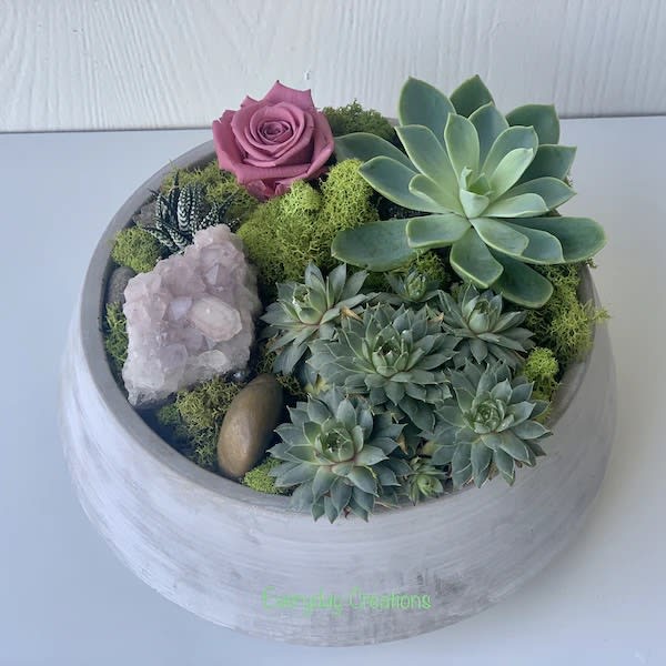 EC06 - Chic Succulent Garden  - Bring a touch of modern elegance and natural beauty into your home or office with the Chic Succulent Garden arrangement. This beautiful garden features a variety of stunning succulent plants that showcase a range of colors, shapes and textures. The succulents are artfully arranged in a stylish cement planter with preserved roses and a rose quartz rock adding a touch of softness and sophistication. The Chic Succulent Garden is the perfect way to add a touch of organic beauty, whether you're looking for a stylish way to spruce up your home decor, or want a unique and thoughtful gift for a loved one, this garden is easy to care for and sure to impress. 
