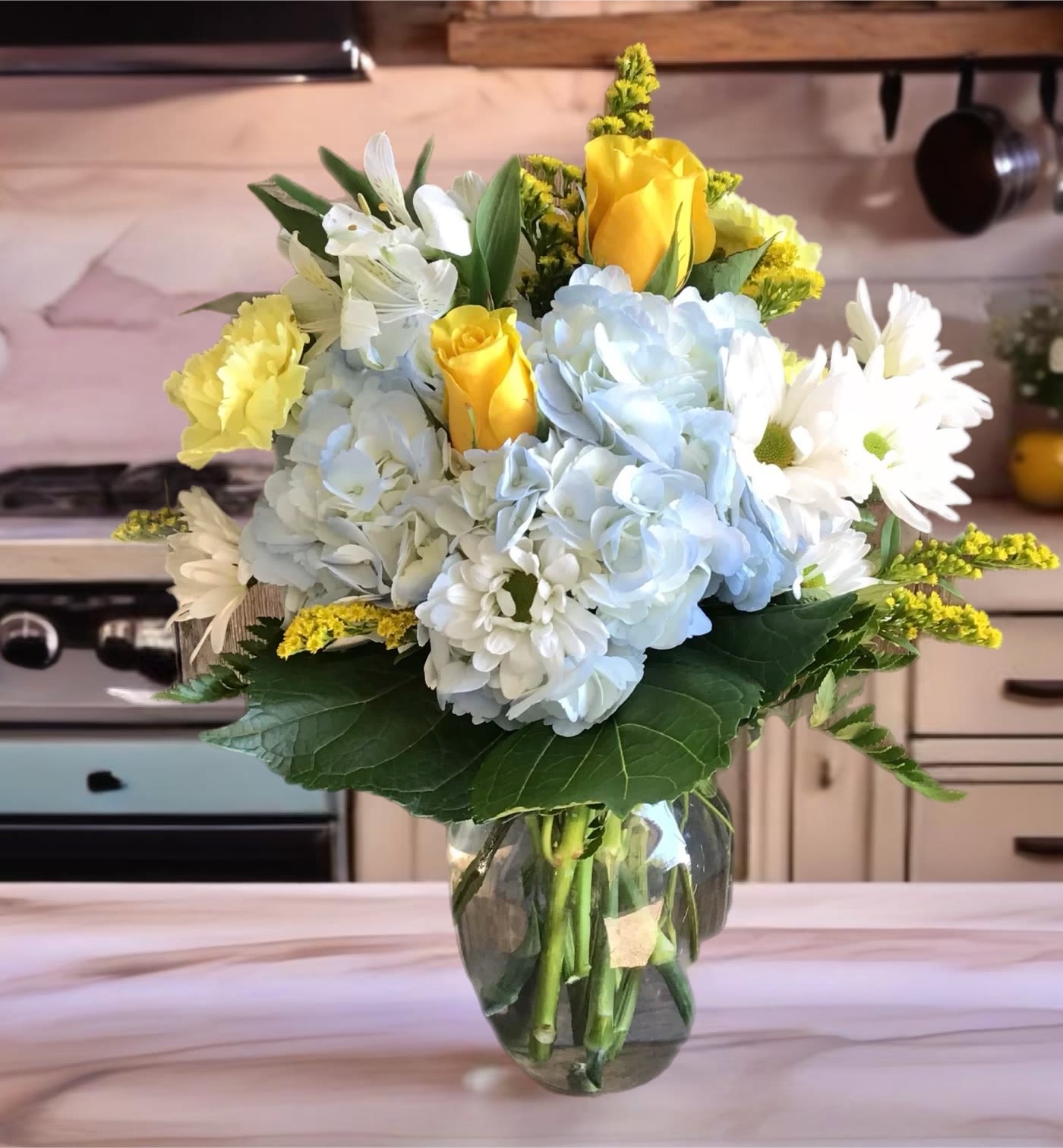 Bouquet of yellow roses, blue hydrangeas, and white daisies in a glass vase