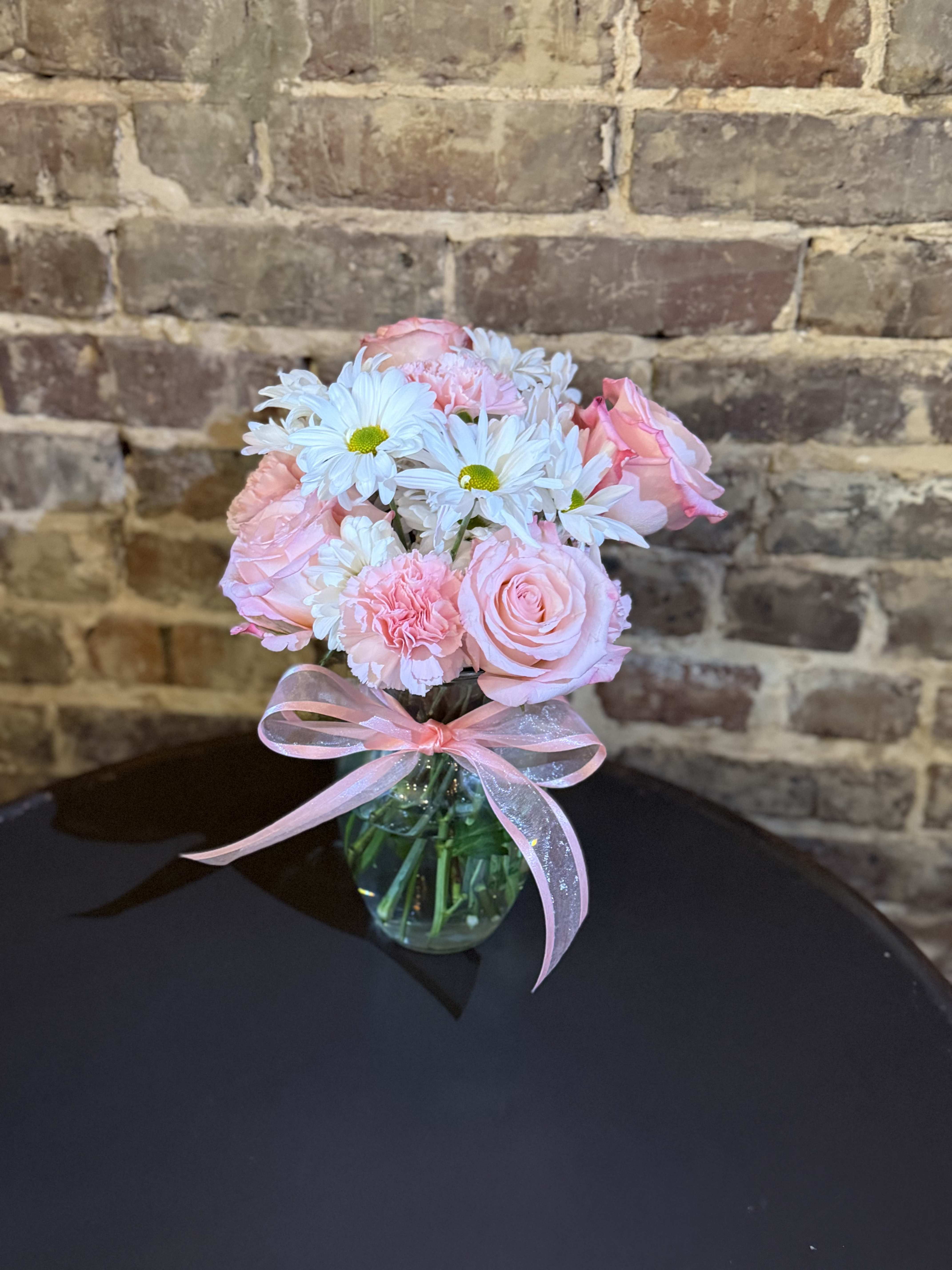 Mother's Day Pink Posies - Soft pale pink roses and ruffled pink carnations mingle with cheerful white daisy-like blooms in a compact, rounded bouquet made for Mother's Day. The gentle pastel palette and delicate textures give the person you're thinking of an immediate sense of being cherished-soft, romantic, and quietly celebratory.  Arranged in a clear glass vase and finished with a sheer pink ribbon, this small arrangement feels gift-ready the moment it arrives. The mix of roses, carnations, and daisies creates varied texture and lasting visual interest while staying sweet and feminine in pink, white, and green tones. Its modest size makes it perfect for a bedside table, kitchen counter, or desk where your recipient will see it often.  Choose this bouquet to say "thank you," "I love you," or "thinking of you" with a floral keepsake that feels intimate rather than grand. Standard care-fresh water in the vase, a cool spot out of direct sun, and a trim to stems if you transfer flowers-will help the blooms stay lovely for days.