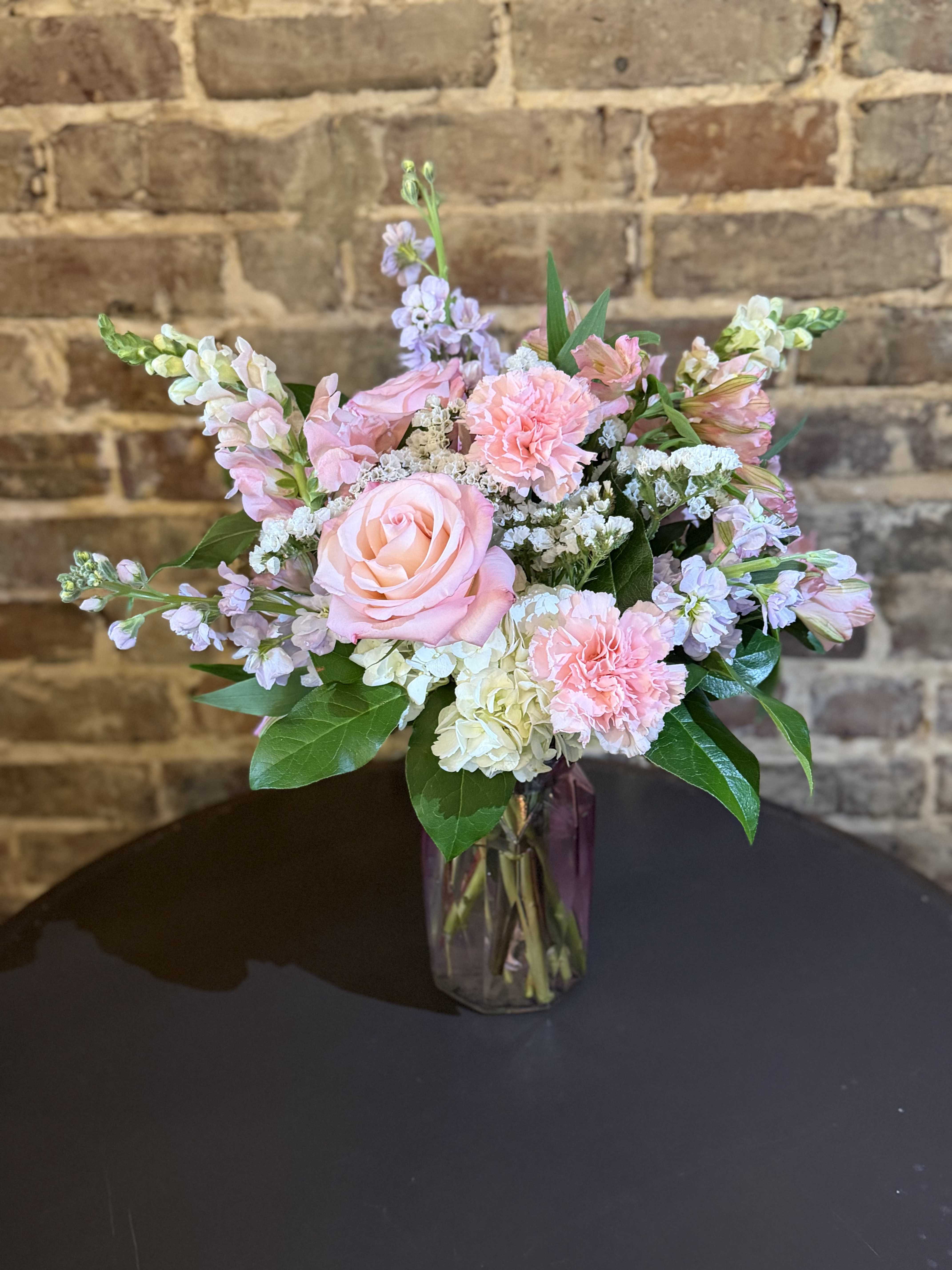 Mother's Day in Pink - A soft pastel bouquet that reads like a garden memory-this Mother's Day in Pink arrangement is a Mother's Day designed to make her feel cherished. Blush roses and pink carnations create a warm, affectionate center while white hydrangea and lavender stock add cloud-like fullness and a delicate, perfume-tinged lift; the overall effect is romantic and gently feminine, perfect for celebrating moms.  Flowers are loosely arranged with airy height and a rounded silhouette in a pink vase, giving it a fresh, natural look that suits a bedside table or dining-room centerpiece. The pink, white, and green palette layers soft textures-velvety rose petals, ruffled carnations, fluffy hydrangea clusters, and elegant stock stems-for visual interest and a light, garden-fresh scent.  Send this to the person you're thinking of when you want them to feel truly seen and loved; it reads as thoughtfully celebratory without being overdone. Arrives ready to display in the included glass vase-trim stems and refresh water every 2-3 days to keep blooms at their best.