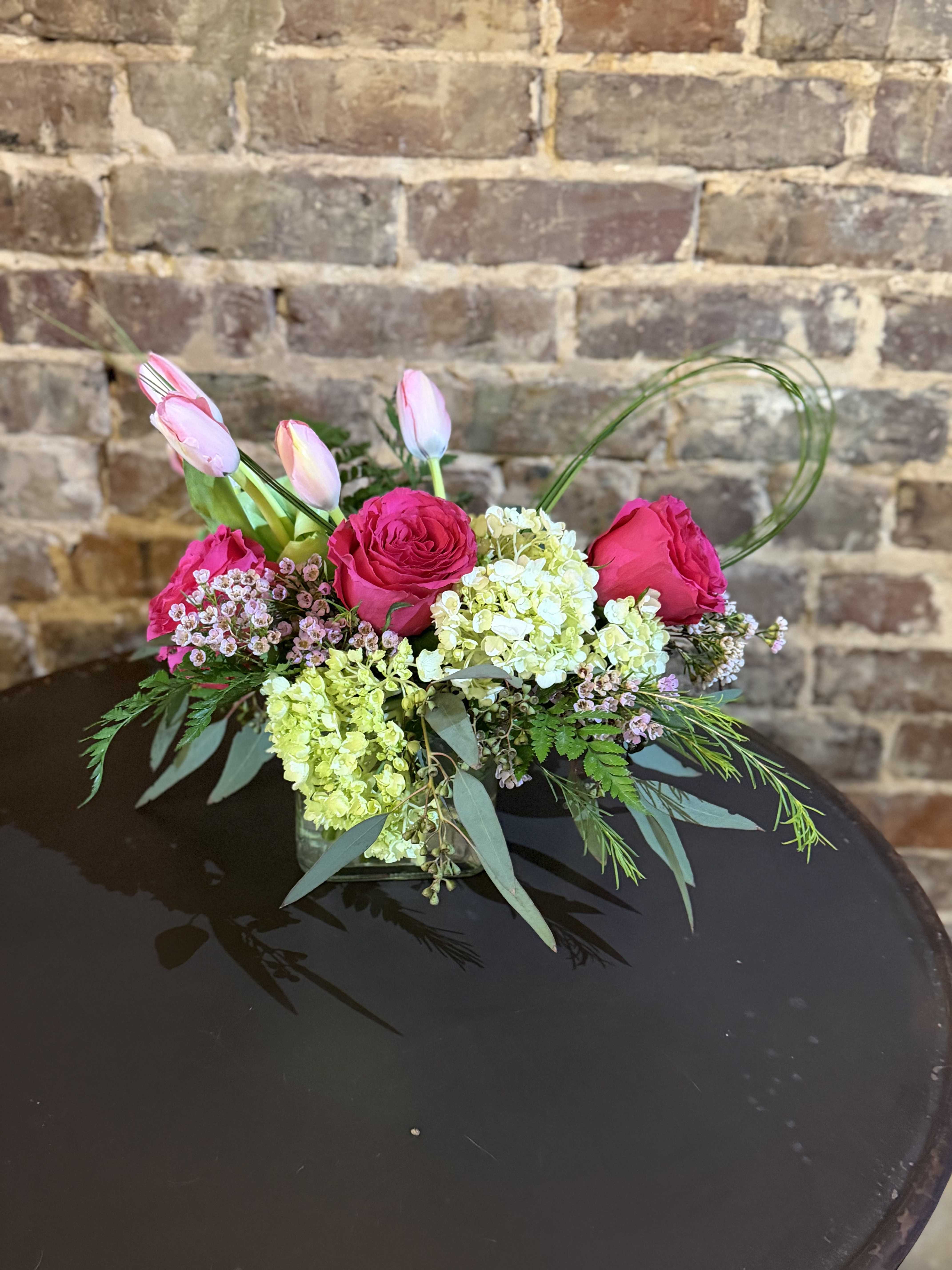 A Mother's Joy - Vivid pink roses paired with pale pink tulips create a bright, cheerful bloom that feels like a warm hug-perfect as a Mother's Day bouquet. This low flower arrangement announces itself with a fresh, slightly romantic tone that will make the person you're thinking of feel seen and celebrated.  Clusters of green hydrangea add lush volume, while small blush filler flowers and airy greenery soften the edges for a natural, garden-fresh look. Arranged compactly in a clear glass vase, the horizontal silhouette is modern and low enough to sit on a dining table or desk without blocking conversation.  A Mother's Joy is designed for everyday gifting as much as special moments-she'll feel genuinely appreciated whether this arrives for a thank-you, a milestone, or just because. The clear vase keeps care simple: fresh water and a clean vase help these blooms last, and the arrangement's low profile makes placement and enjoyment effortless.