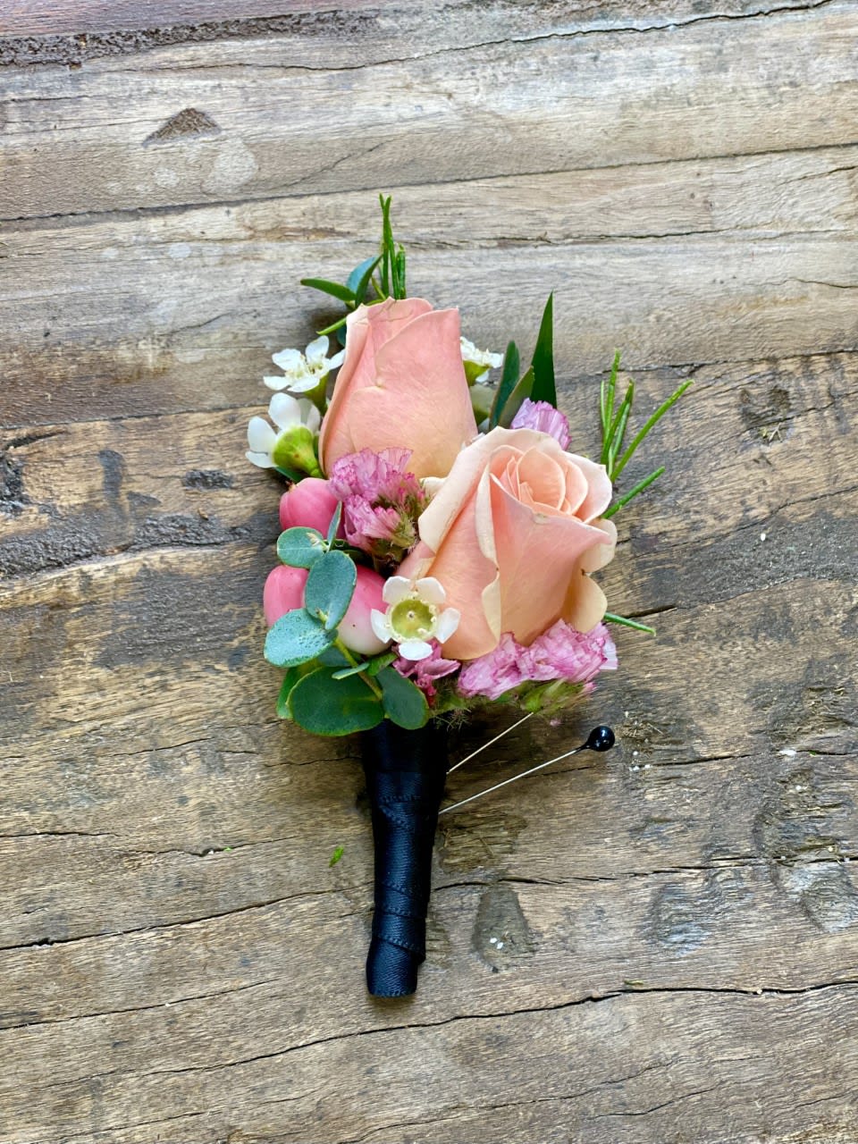 Peach rose boutonniere with pink and white accent flowers wrapped in black ribbon on rustic wood.