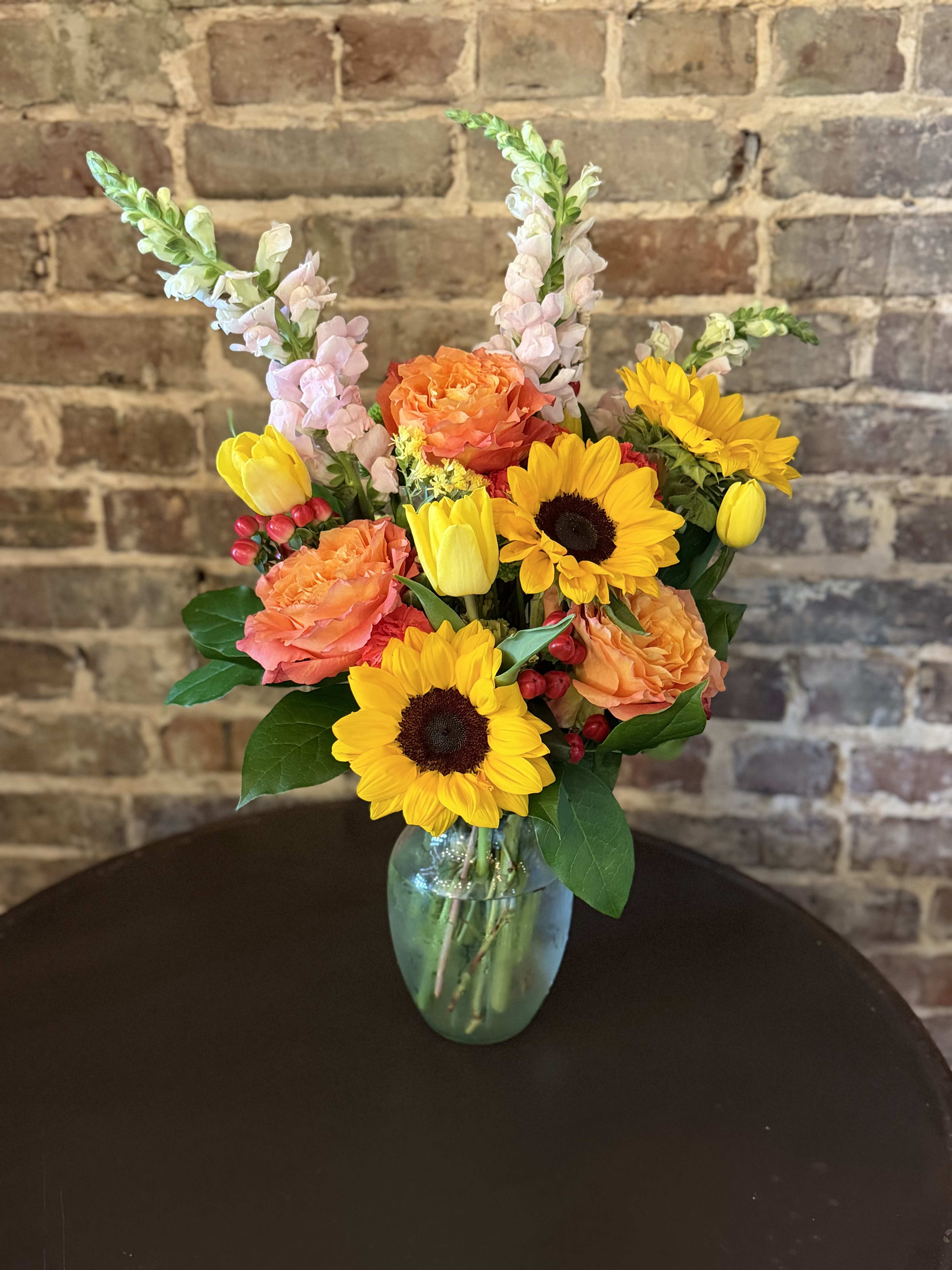 A Mother's Love - Bright, garden-fresh sunflowers take center stage in this cheerful bouquet, paired with orange roses and sunny yellow tulips for a warm, celebratory feel perfect for Mother's Day. The arrangement is designed to make the person you're thinking of feel noticed and adored-the bold yellows and oranges read as joy and gratitude, while pale pink snapdragons add a soft, thoughtful note.  The arrangement features bright yellow sunflowers, orange roses, yellow tulips, and pale pink snapdragon spikes artfully arranged in a clear glass vase. The composition is full and upright: taller snapdragons rise above the rounder sunflowers and roses for a layered, garden-style silhouette that reads fresh and seasonal on a table or mantle. The clear glass vase keeps the focus on the vivid blooms and lets the stems show through for a clean, classic presentation.  A Mother's Love is the kind of bouquet that makes someone feel genuinely celebrated-ideal for moms, mentors, or anyone who brightens your life. Vase-ready and easy to display, it arrives arranged and hydrated; for best care, refresh the water every two days and trim stems slightly to prolong bloom life.
