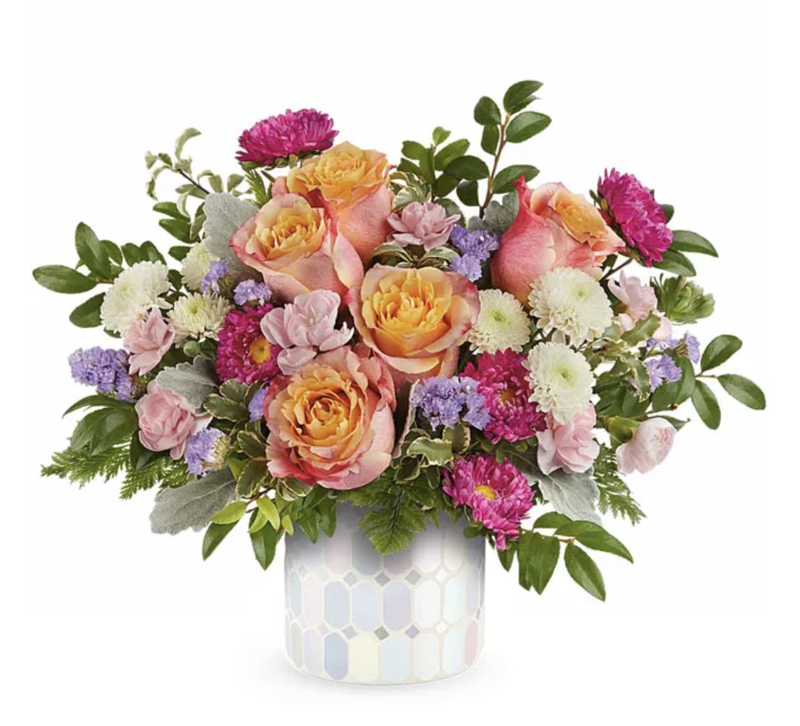 Blooming Iridescent Arrangement - Brighten someone's day with this charming floral arrangement, designed to bring warmth and joy to any occasion. Soft light orange roses take center stage, beautifully complemented by delicate miniature pink carnations. Hot pink Matsumoto asters add a vibrant touch, while crisp white button spray chrysanthemums provide contrast and freshness. Accents of lavender statice bring a hint of elegance, and lush floral greenery ties it all together. Arranged in a stylish pastel hexagon-patterned ceramic container, this bouquet is a thoughtful and uplifting gift for any celebration.