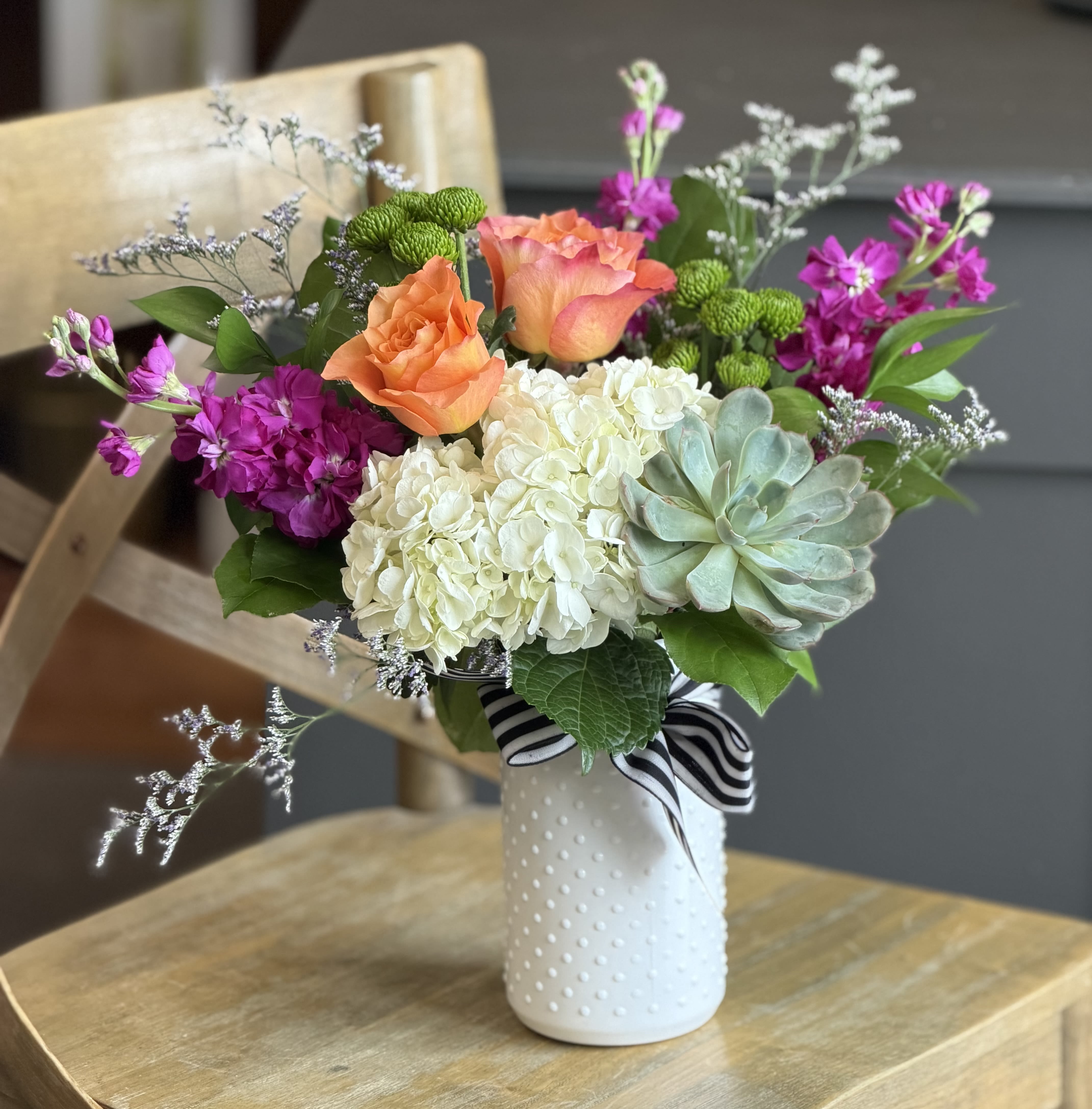 Sunset Afterglow - A luminous mix of orange roses, white hydrangea, magenta stock and a pale green succulent creates an unforgettable bouquet perfect for an anniversary, birthday, Mother's Day, or a "just because" surprise. This flower arrangement delivers a warm, romantic glow-she'll feel noticed and cherished the moment it arrives-while the airy branching accents add movement and a light, celebratory silhouette.  The design is full and rounded, centered on a large white hydrangea  surrounded by vibrant orange roses, bright magenta stocks (and other seasonal fillers), and a soft succulent that adds texture and modern contrast. It comes in a white textured ceramic vase finished with a black-and-white striped ribbon for a polished yet playful touch. Average dimensions fit nicely on a dining table or desk; vase and stems are arranged for immediate display with simple care: fresh water and a cool spot will keep blooms looking their best.  Ideal for expressing love and romance or marking a milestone birthday, this arrangement also reads as an effortlessly thoughtful everyday gift-bright, tactile, and ready to make someone feel truly seen. Order with confidence for local delivery and same-week pickup when available.