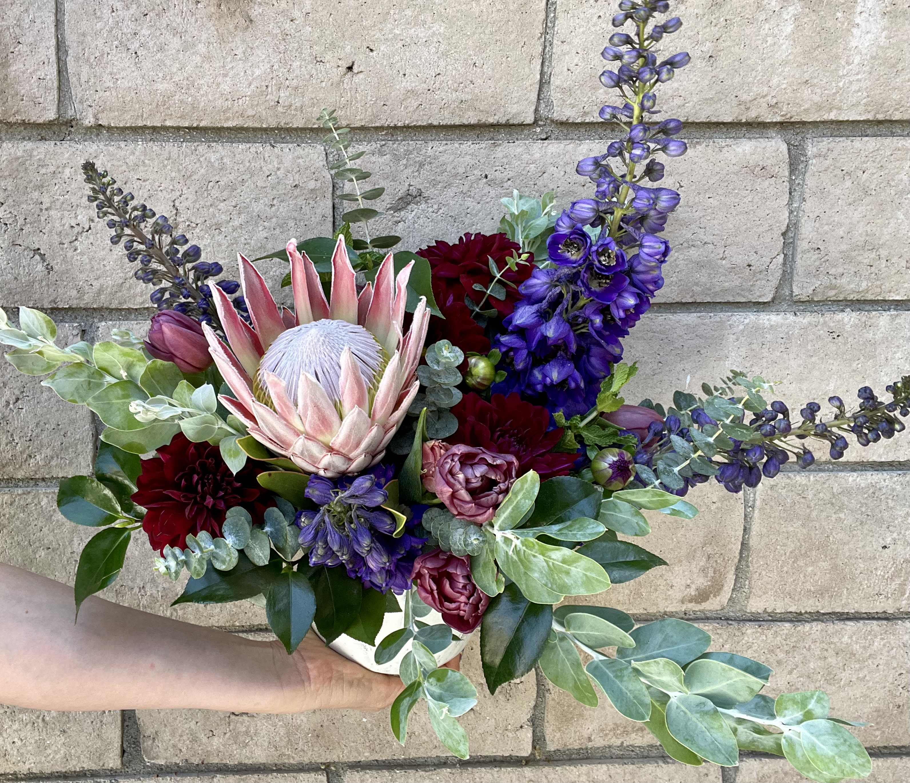 Queen Bee - A divine gift where the Queen Protea is the star. Nestled in with blue Hybrid Delphinium, purple Tulips and burgundy Dahlias- this gift will take their breath away. 