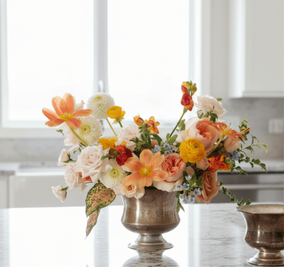 Low centerpiece of peach, yellow, and white flowers in a silver compote vase on a kitchen counter
