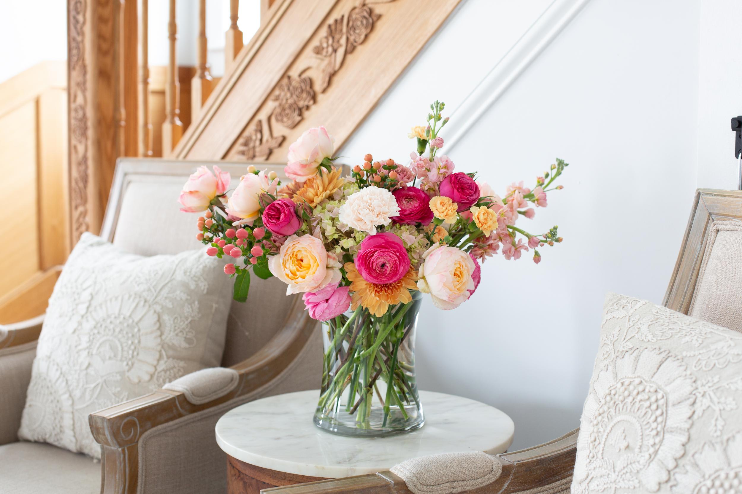 Mixed bouquet of pink, peach, and yellow flowers in a clear vase on a small table between chairs