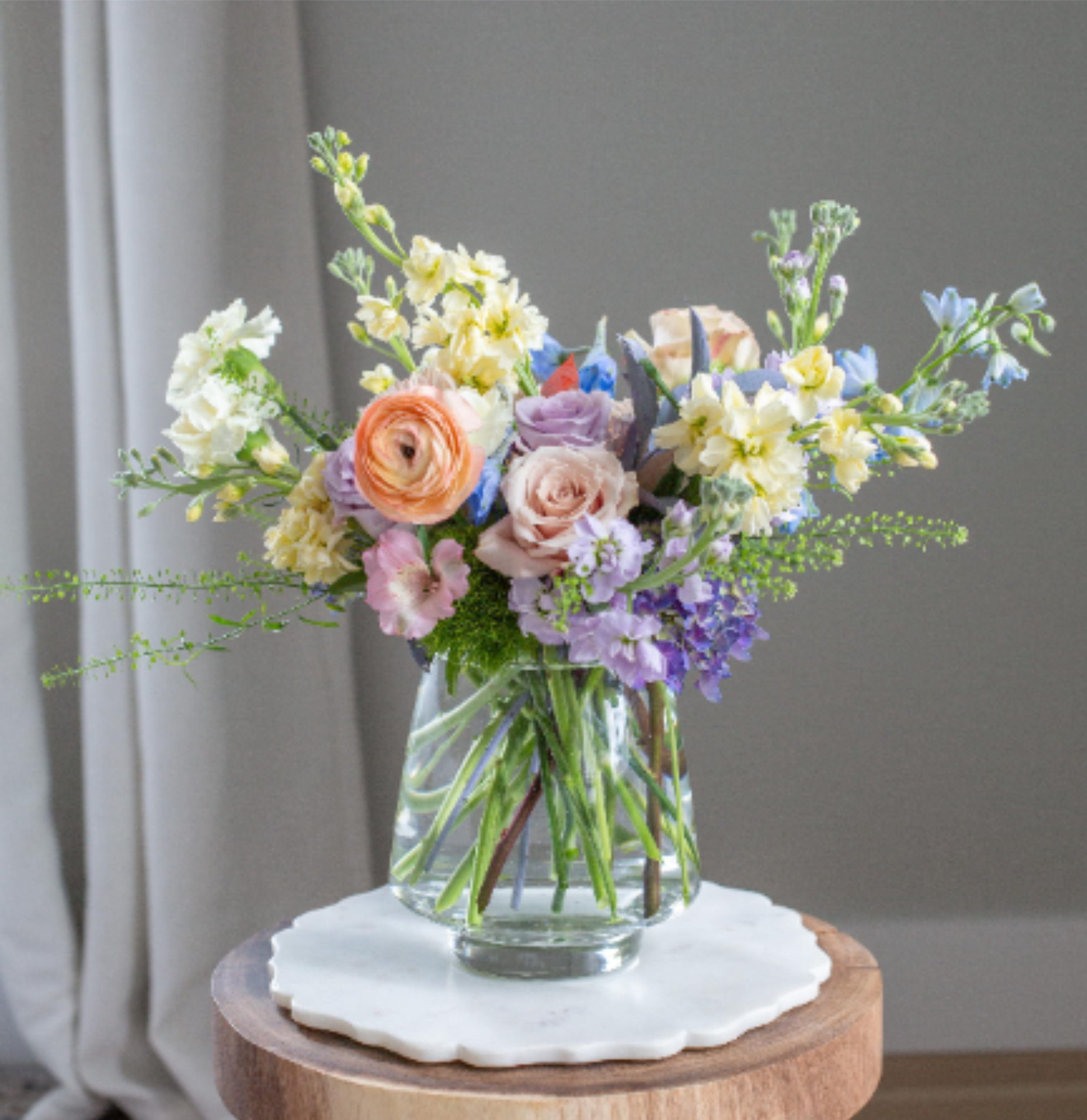 What a Wonderful World – Spring Garden Arrangement - Inspired by the timeless song made famous by Louis Armstrong, What a Wonderful World celebrates the beauty, warmth, and gratitude we feel for the people who make life brighter.  This airy spring garden arrangement features a delicate palette of soft lavender, butter yellow, peach, and fresh spring greens. Seasonal blooms such as ranunculus, garden roses, stock, delphinium, and textural spring flowers are arranged with natural movement in a modern clear glass vase.  The design feels light, organic, and naturally elegant capturing the feeling of a blooming spring garden. Soft lavender and blue tones add gentle color while peach and butter yellow blooms bring warmth and brightness to the composition.  Each arrangement is handcrafted by the designers at ImpeKable Flowers in DeLand, using carefully selected seasonal flowers sourced from premium growers.  A beautiful choice for Mother’s Day, birthdays, thank-you gifts, or celebrating someone who makes the world feel a little more wonderful.  We proudly offer flower delivery throughout DeLand, Lake Mary, Sanford, Deltona, Orange City, DeBary, Altamonte Springs, and surrounding Central Florida communities.  Flower varieties may vary slightly based on seasonal availability while maintaining the same color palette and airy garden style.