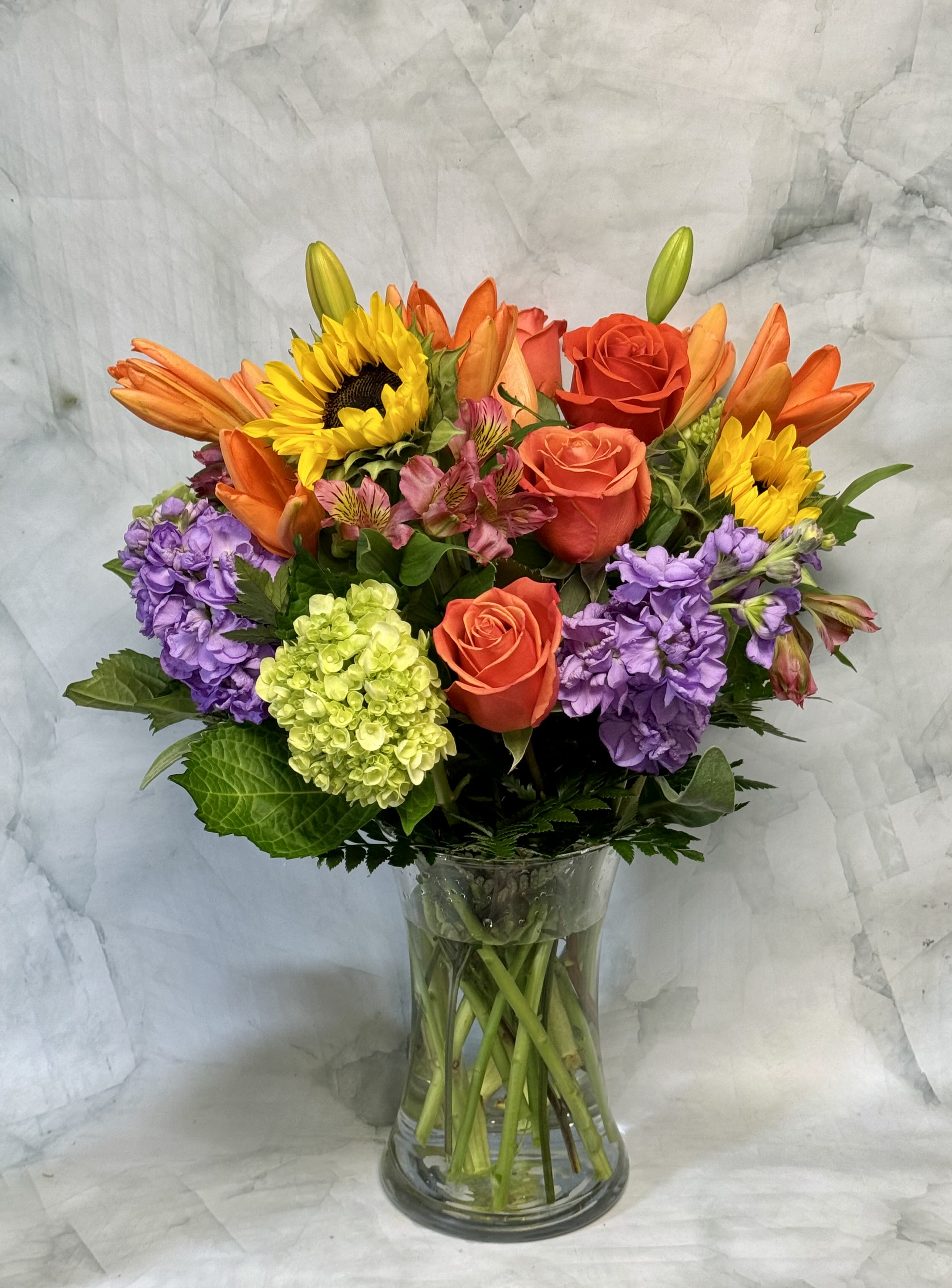 Mixed bouquet of roses, sunflowers, lilies, and purple blooms in a glass vase