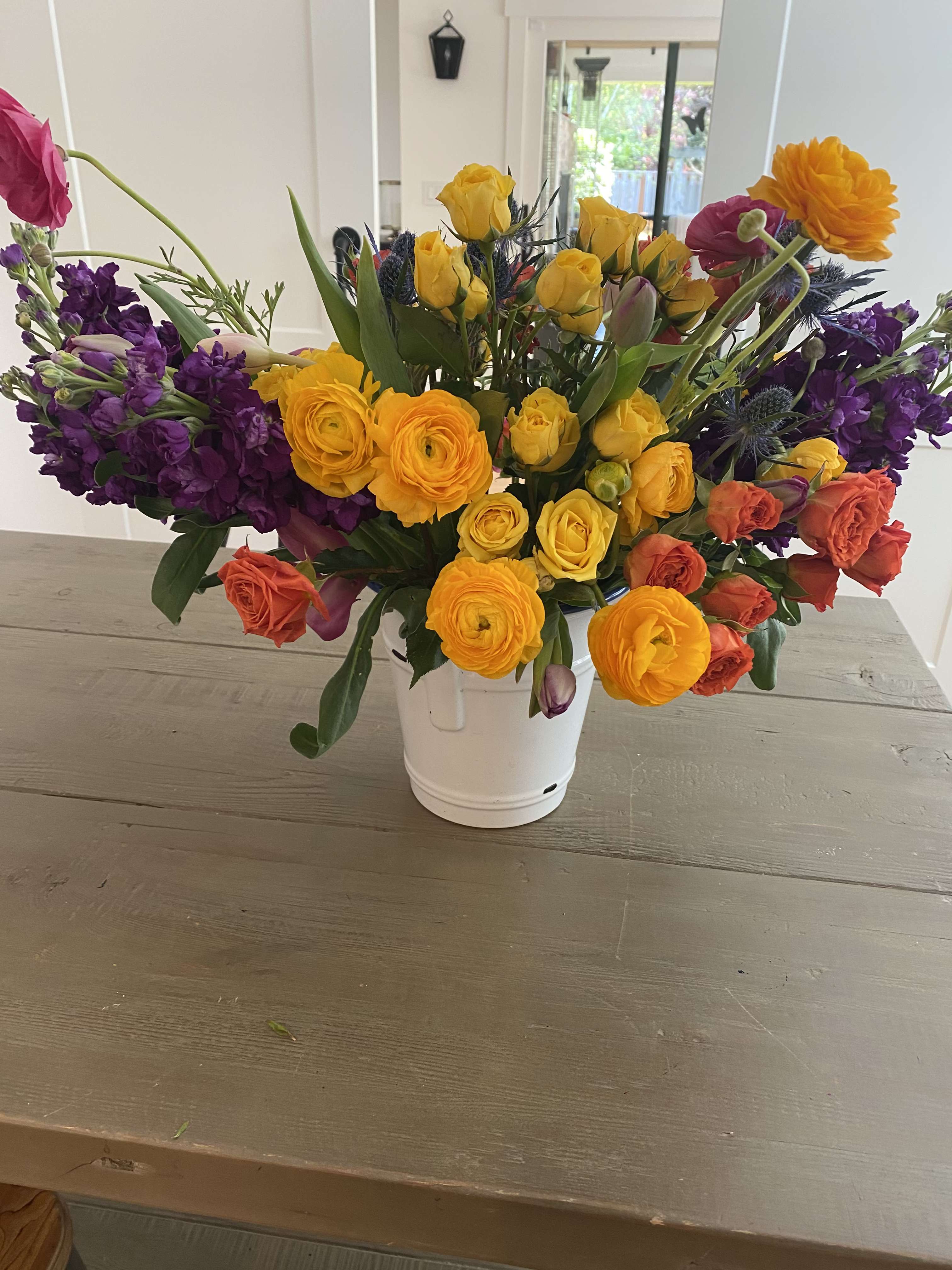 Colorful mixed bouquet in a white bucket vase with yellow, orange, pink, and purple blooms.