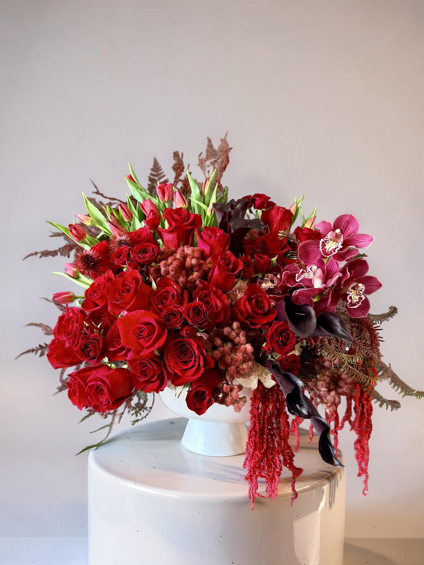 Ruby Velvet  - A lush, sculptural bouquet of deep red roses and rosebuds set against pink tulips and magenta orchids - Ruby Velvet is a romantic flower arrangement perfect for an anniversary, Valentine's Day, or any moment you want someone to feel deeply seen. The dramatic crimson palette and trailing red amaranthus give this arrangement a luxurious, formal presence that also makes a striking centerpiece for holiday dinners, graduations, or a bold birthday gift.  This dense, low-to-medium composition pairs dark purple calla lilies and burgundy accents with fern-like greenery for texture and movement, assembled in a clean white pedestal vase that elevates the arrangement's sculptural silhouette. The asymmetrical layers and cascading elements create visual flow from every angle, making it ideal as a mantlepiece display or dining-table focal point.  Give Ruby Velvet to the person you're thinking of when you want to convey love, admiration, or sympathy with elegance - it's equally fitting for Mother's Day, Memorial Day, Christmas, or a "just because" surprise. Designed to arrive full and ready to display, this arrangement suits formal and celebratory settings; refresh water as needed and place away from direct heat to keep blooms looking their best.