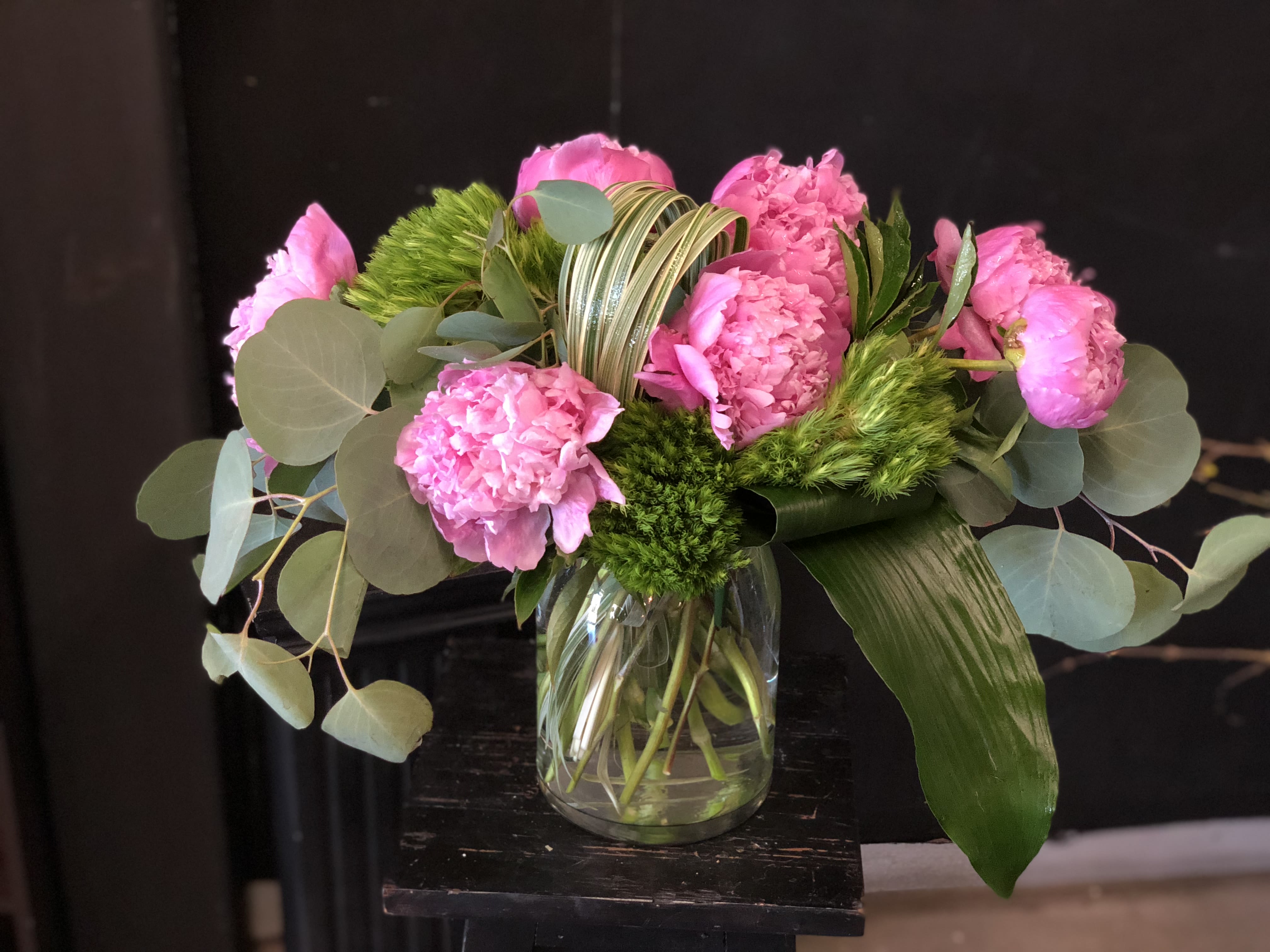 Peony Delight - Clustered pink peonies take center stage in this cheerful birthday bouquet, their ruffled petals softened by sprigs of textured green filler and broad, round eucalyptus leaves-an ideal choice for birthdays, thank-you gifts, or spring celebrations. The arrangement reads romantic without being fussy, the striped decorative loop and a large glossy leaf adding modern structure and contrast so the person you're thinking of feels both cherished and delighted.  Arranged in a clear glass vase that shows the airy, hand-tied stems, the mix of pink, green, and white tones keeps the palette fresh and versatile. Carnations provide long-lasting color and a soft, full silhouette while the eucalyptus brings subtle scent and sculptural shape; decorative accents highlight the bouquet's contemporary lines. This size works well on a bedside table or small dining centerpiece; stems are visible through the vase so care is simple-fresh water and a quick trim every few days will help the blooms last.