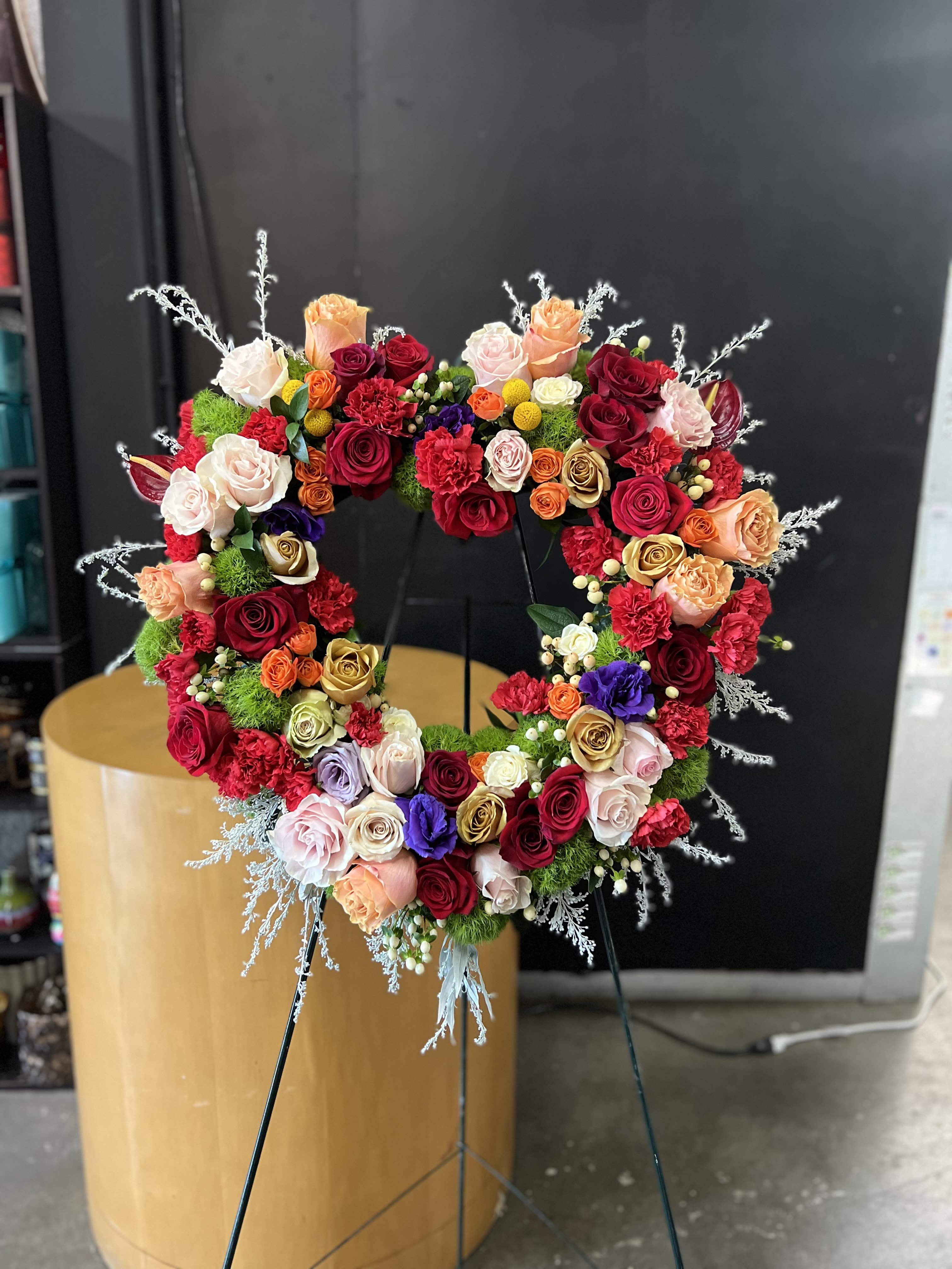 Heart Shape Celebrating Life - A lush heart-shaped wreath of roses and carnations creates an immediate, emotive statement-perfect for a sympathy tribute or a heartfelt display at a memorial or celebration of life. The dense heart silhouette reads clearly from a distance, so the person you're honoring will feel deeply remembered and the space will carry a warm, loving presence.  This arrangement is packed with roses in deep red, blush, peach, cream and gold tones with a few purple accents, interspersed with red carnations and small berry-like fillers for texture. Airy silver-white branches extend from the edges to add contrast and a touch of lightness against the rich, layered blooms. Mounted on a sturdy floral stand, the wreath is finished with decorative berries and arranged densely for a dramatic, formal presentation.  Suitable for a funeral, memorial service, or as a long-lasting display to honor someone special, this heart wreath communicates love, remembrance, and celebration. For placement, the included floral stand ensures the piece is prominently displayed; please specify delivery details and any personalization requests when ordering so we can prepare the arrangement to best suit the service.