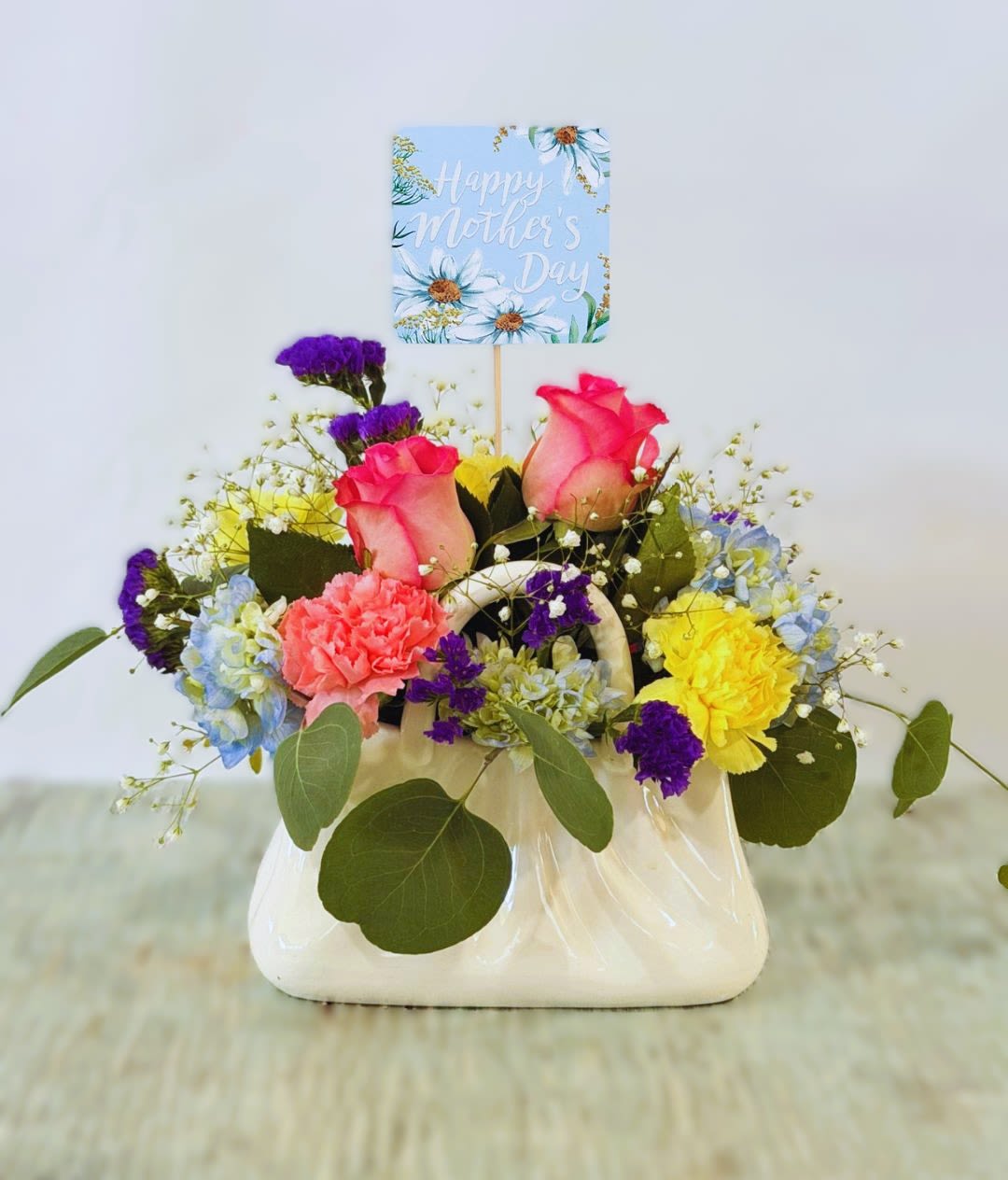 Mother's Day #6 - A playful cluster of pink roses and pale blue hydrangeas arranged in a glossy white ceramic purse-shaped container makes this Mother's Day flower arrangement an instantly cheerful gift. Designed as a Mother's Day bouquet, it reads as warm and celebratory-the printed "Happy Mother's Day" card pick rising above the blooms ensures the recipient knows this was chosen with family and appreciation in mind.  Bright yellow and coral carnations add sunny contrast to the soft pink roses and hydrangea, while small white and purple accent flowers fill the compact, hand-held design. The ceramic handbag vase gives the arrangement a charming keepsake quality and sits neatly on a table or countertop; the size is ideal for a bedside or kitchen display without overwhelming the space.  Perfect for moms who love fresh, colorful florals, this arrangement says "you're seen and loved" whether it's for Mother's Day, a birthday, or a thank-you gesture. Lightly mist the blooms and change the water every couple of days to help them last longer; the vase is ready to display as soon as it arrives.