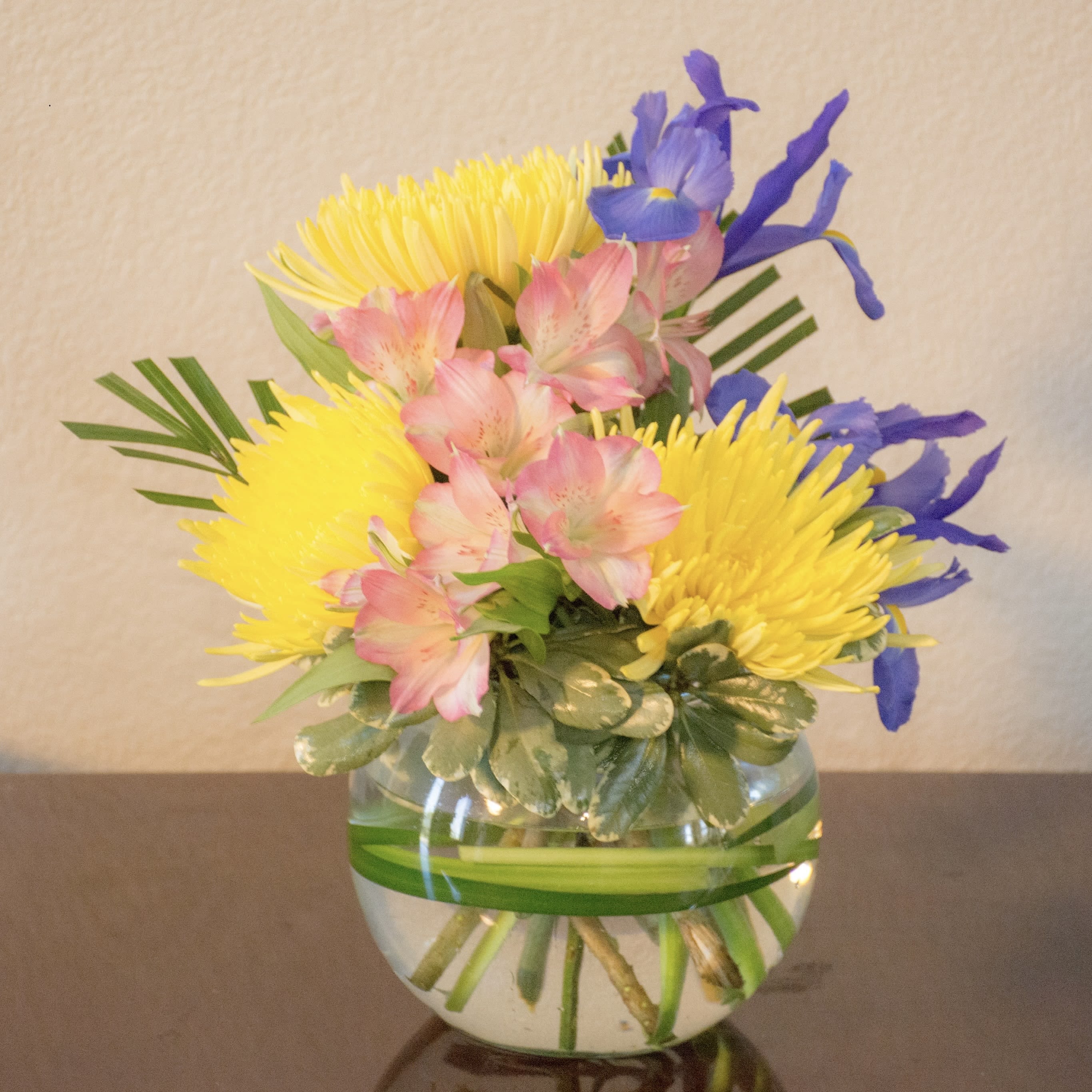 Small arrangement of yellow, pink, and blue flowers in a round glass vase with a green leaf wrap.