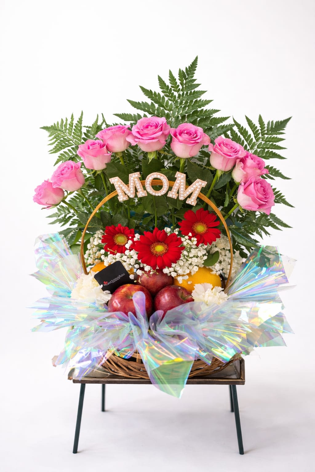 Mom’s Garden Delight Basket - A ring of soft pink roses encircling bright red gerbera daisies makes a joyful, celebratory statement-perfect as a Mother's Day flower arrangement to make the mom in your life feel honored and adored. The mixed color palette of pink, red and white reads both classic and festive, so it also works beautifully for birthdays, thank-you gifts, or "just because" moments when you want her to feel truly seen.  This woven basket design layers clustered white filler blooms with roses and gerberas for a textured, abundant look. Fresh fruit is tucked into the lower portion of the basket for a gift-basket feel rather than a standard vase display, and an iridescent decorative wrap catches the light. A metal "MOM" topper with jeweled detail and an included gift card complete the presentation, giving the arrangement extra sparkle and a keepsake element.  Give this to the mother, grandmother, or mentor whose warmth you want to celebrate-the kind of gift that reads as thoughtful, festive, and generous. Order with delivery instructions and your personalized message on the included card; the basket arrives ready to display with minimal care beyond fresh water for the flowers and a cool spot out of direct sun.