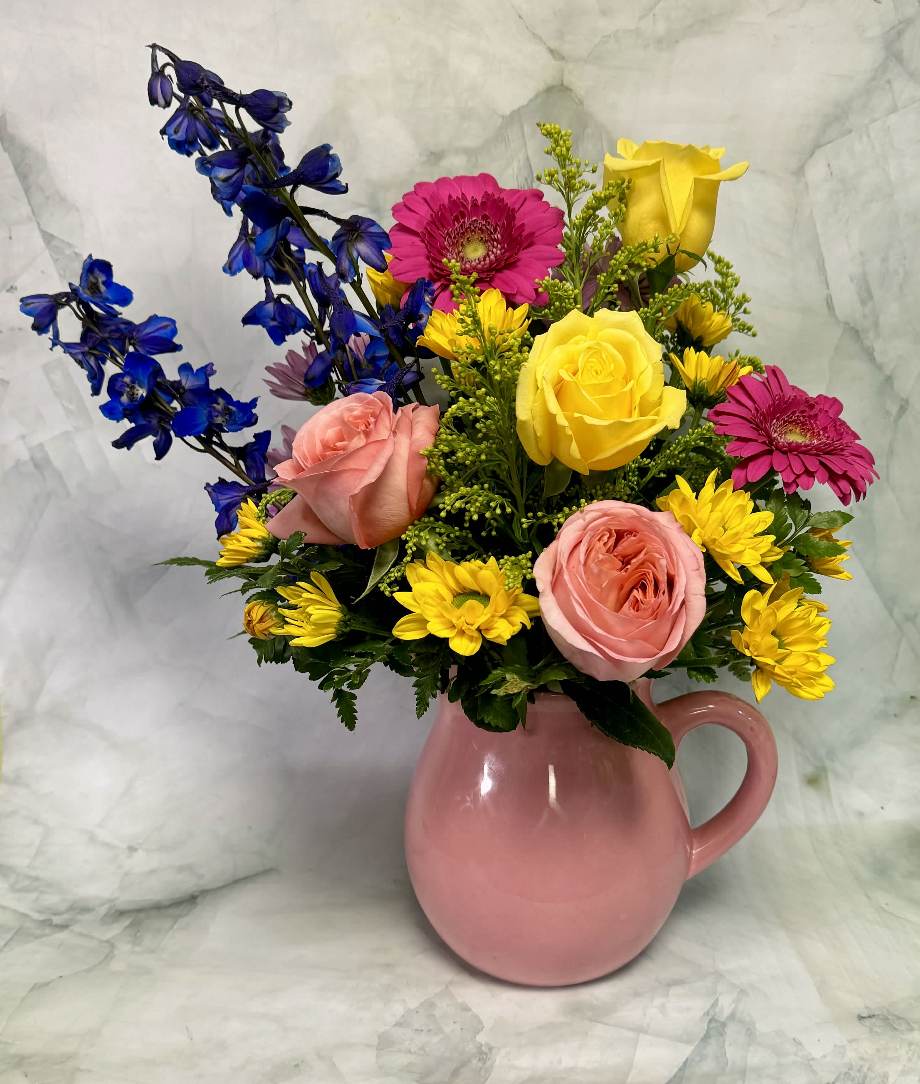 Mixed bouquet of pink, yellow, and blue flowers in a pink pitcher vase