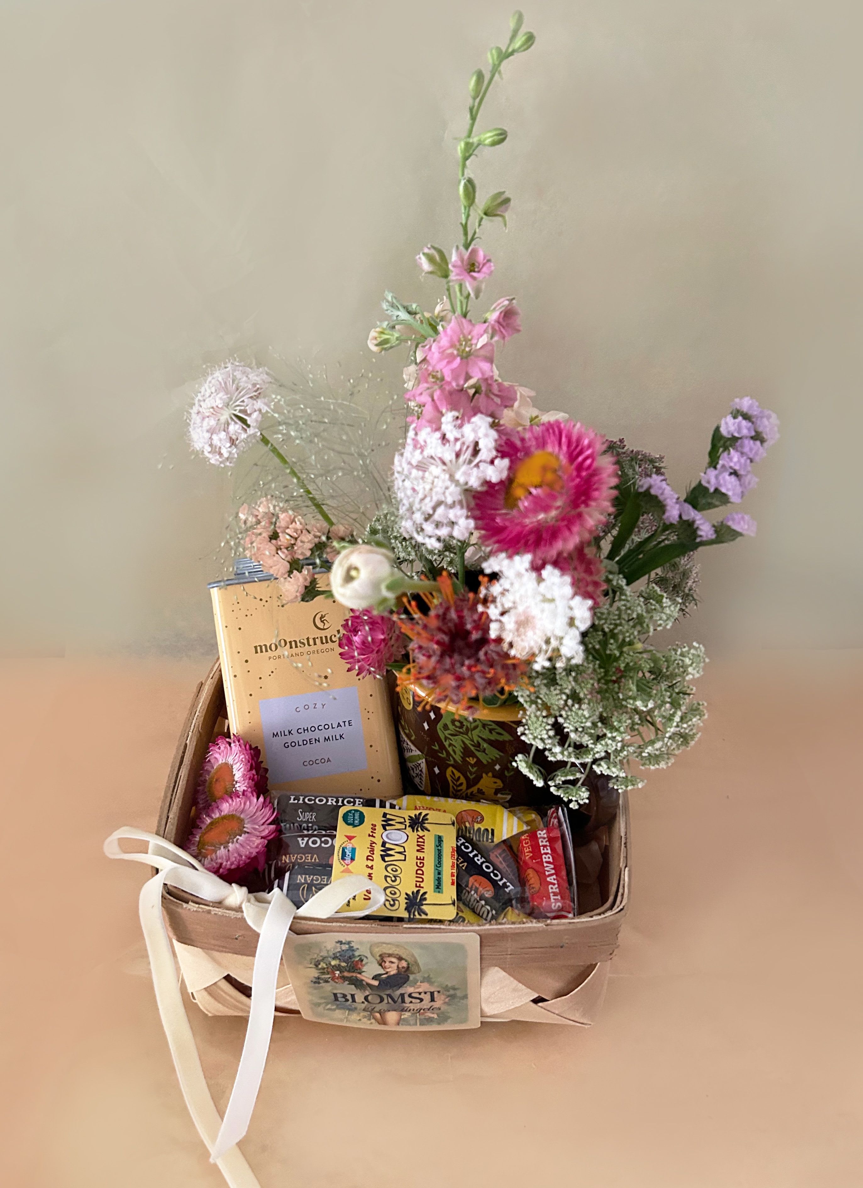 Hygge Basket - A lovely, natural basket in golden colors in the flavors of golden milk and coconut. The petite and classic wooden French basket measures 7" by 7" by 4" and is overflowing with lovely treats and flowers. The basket contains: Enamelware Coffee &amp; Camp Mug, design: Woodland Friends | 16 oz. The cup is illustrated with all the animals of the Forests. Moonstruck Golden Milk Hot Cocoa: Embrace the comfort and strength of the moon with calming ginger and turmeric in milk chocolate. Ingredients: Powdered sugar, Dutch-processed cocoa powder, ginger powder, and turmeric powder. 9.5 Ounces. Coco Wow: Vegan &amp; Dairy Free Assorted Fudges 10oz Bag. Organic, vegan and gluten-free sweets with short ingredient lists and clean labels, they are made only from certified ingredients that come from organic farming, they contain raw materials that are rich in nutrients, vitamins and minerals. Flavors Include: Toffee, Cocoa, Coconut, Salted Caramel, Ginger, Licorice, Strawberry, and Banana. A mini Blomst arrangement of fresh, pretty stems in a clear cylinder vase 4" by 4". Velvet riboon. ready for hand delivery in LA.