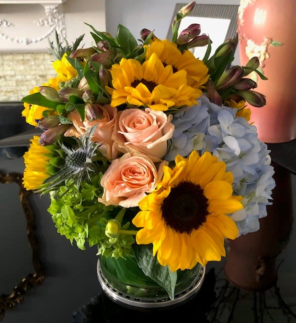 Bouquet of sunflowers, pale roses, blue hydrangea, and green blooms in a glass vase