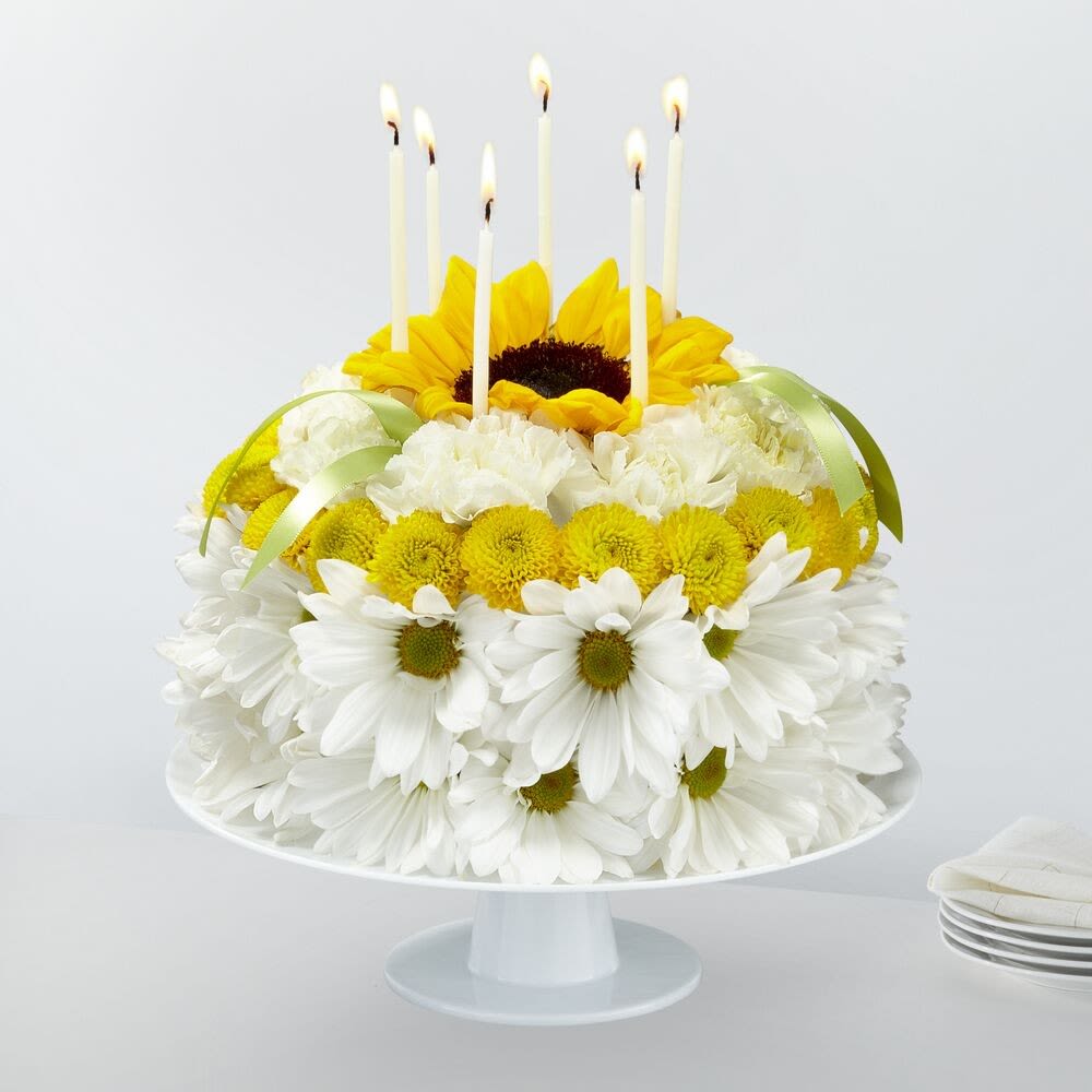 154666 - Blow out the candles, and help make it their brightest birthday yet with our Birthday Smiles Floral Cake. This one–of–a–kind arrangement is frosted with a collection of bright sunflowers, yellow button pompons, white daisy pompons and white carnations. Topped with birthday candles and satin ribbon, this unique gift will bring a smile to their face and make it a truly memorable celebration.