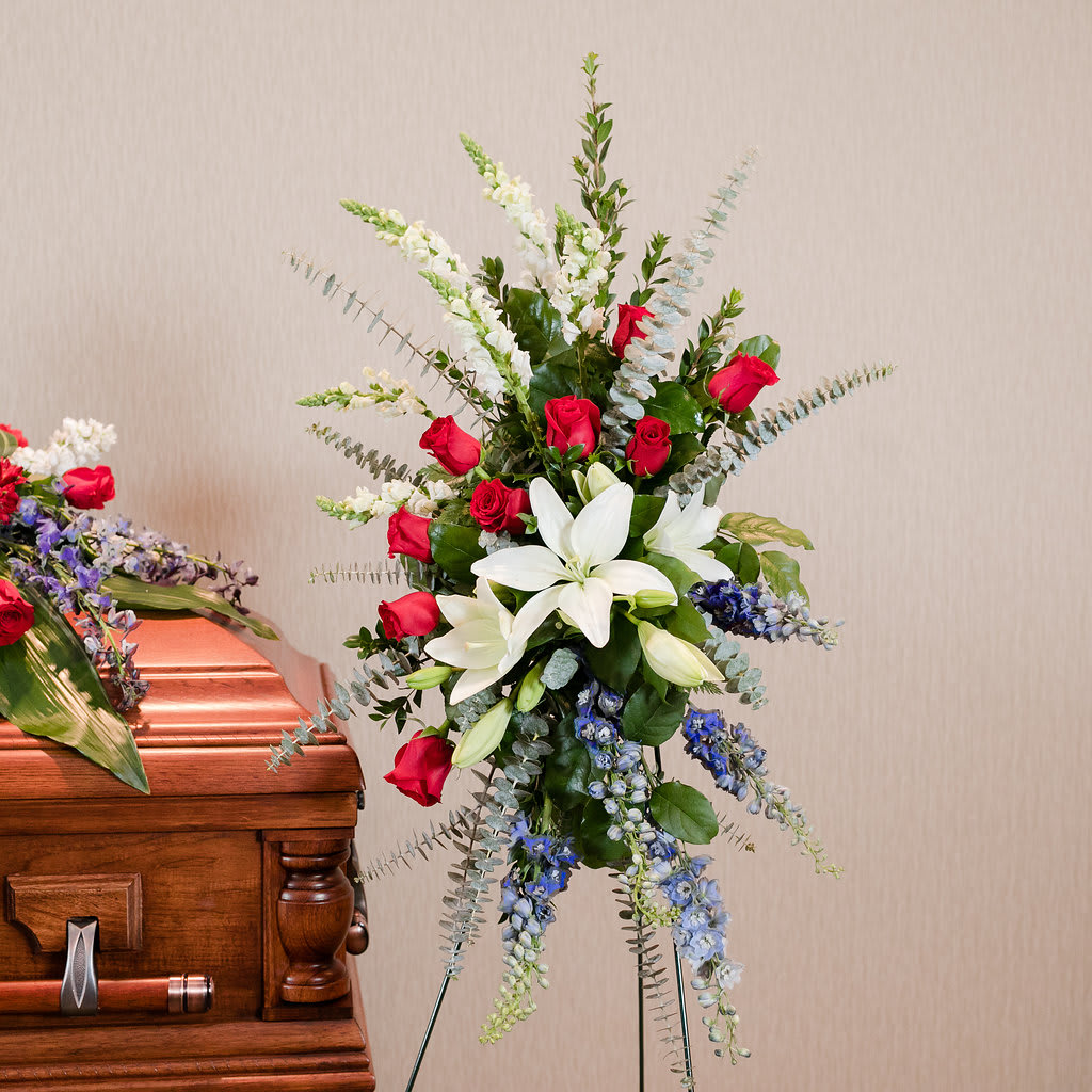 Standing floral spray with red roses, white lilies, and blue flowers beside a casket