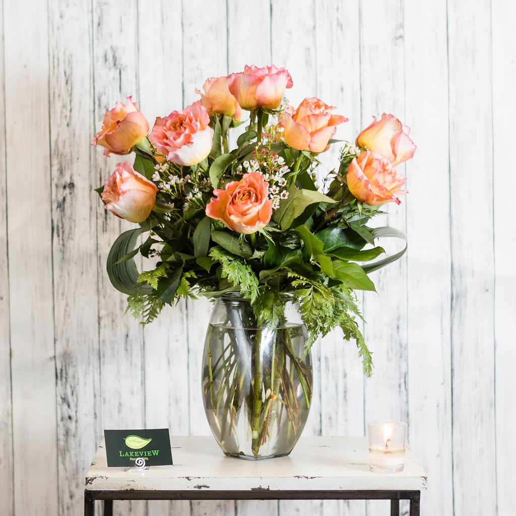 Peach and pink roses with small white blooms arranged in a clear glass vase on a rustic table