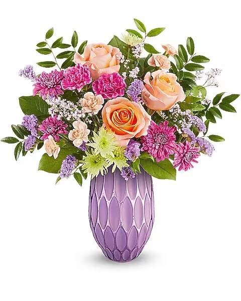 Mother's Glow - Peach roses gathered with bright pink chrysanthemums and sprigs of lavender-hued filler create a warm, romantic Mother's Day or birthday bouquet that feels cheerfully feminine and entirely gift-ready. This flower arrangement for mom blends purple, pink, peach, green, and white tones to make the recipient feel cherished-perfect for anniversaries, thank-you gestures, or simply because you're thinking of her. Order today for same day delivery.  The blooms are densely layered into a full, rounded, garden-style shape with airy greenery extending outward for balance and texture. Finished in a tall lavender glass vase with a textured oval pattern, the piece has a modern, decorative presence on a dining table or bedside. Sized to be noticed without overwhelming a room, it arrives arranged and ready to display; change the water every few days and trim stems to keep the roses and chrysanthemums looking fresh longer.