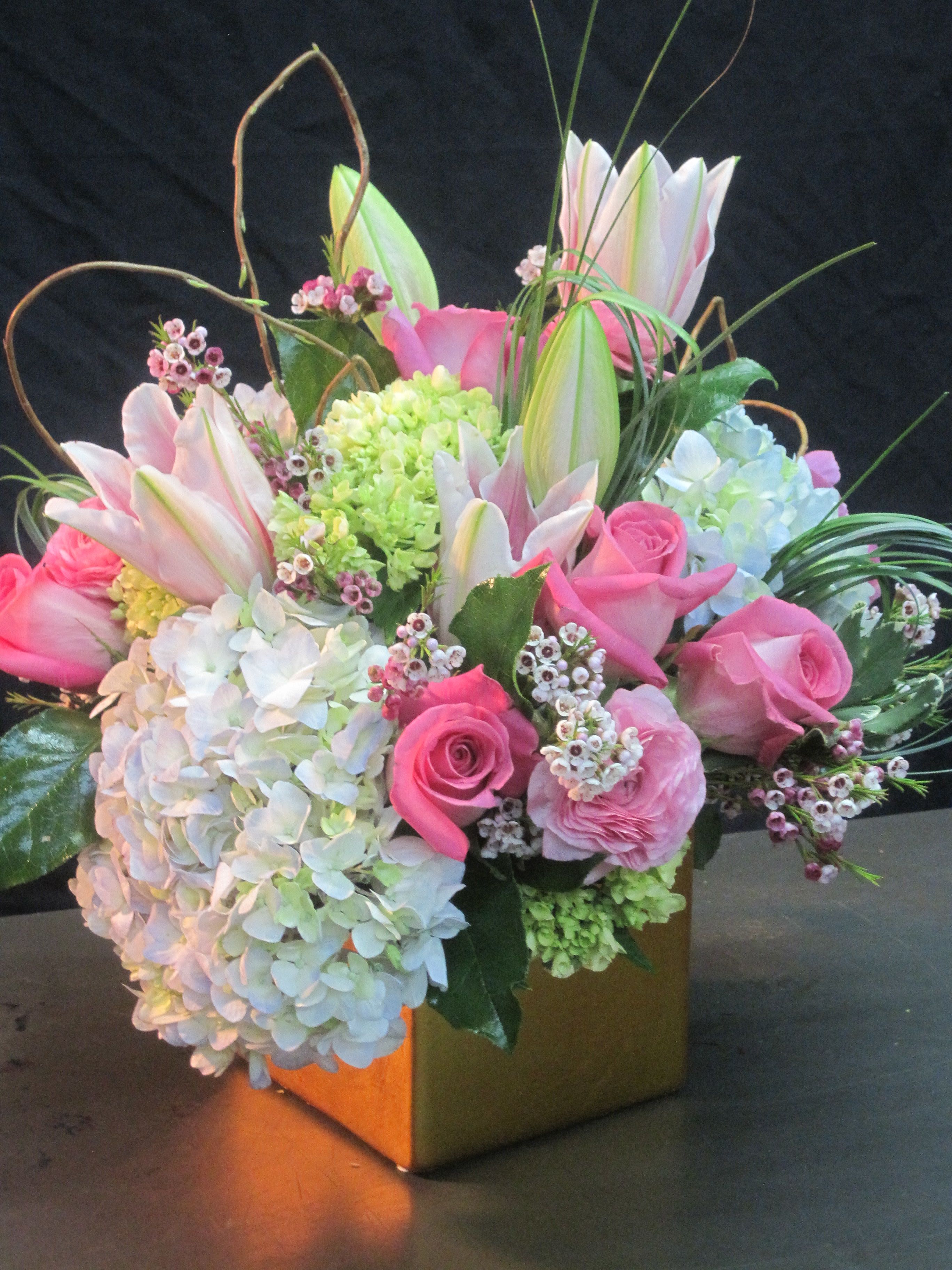 Compact arrangement of pink roses, white hydrangeas, and lilies in a gold cube vase