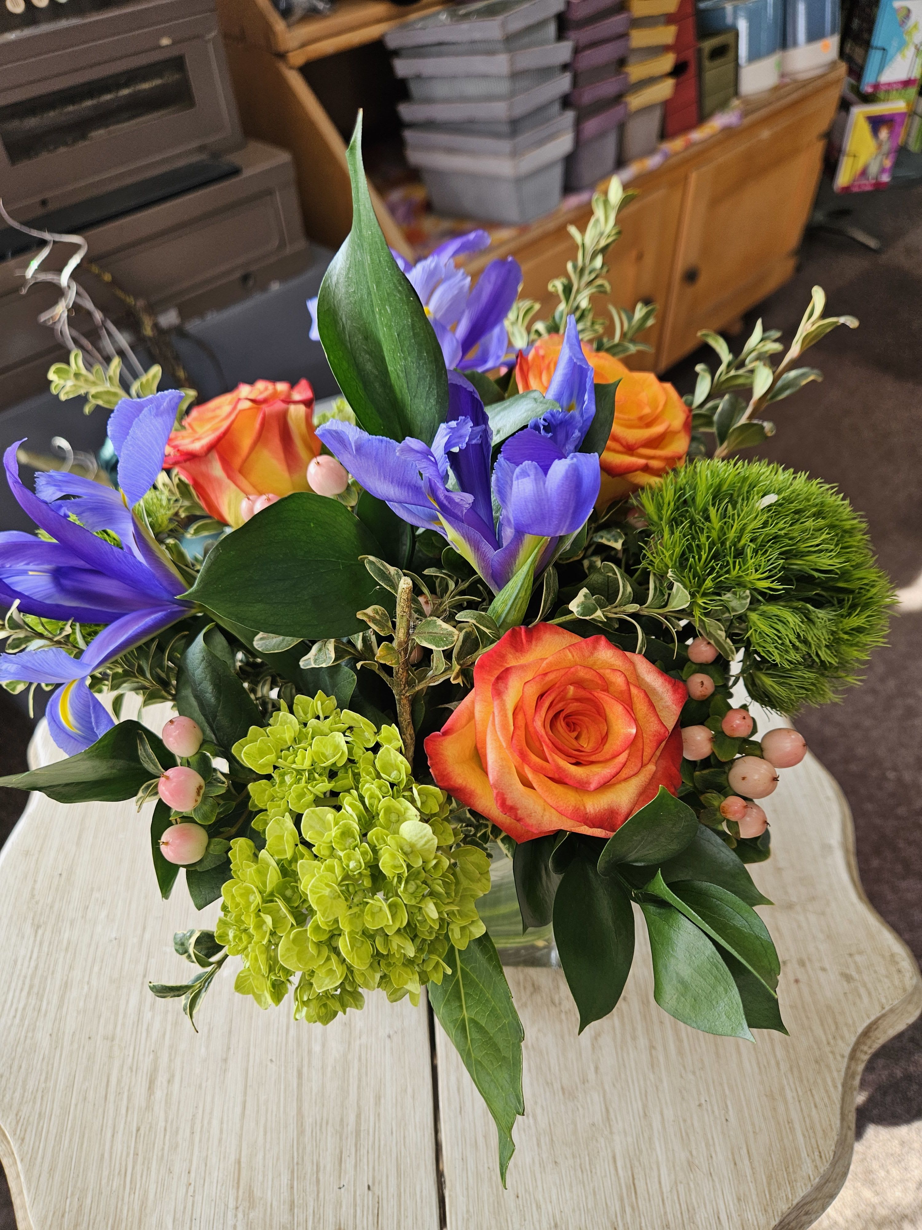 Low arrangement of orange roses, purple irises, and bright green blooms in a clear glass vase