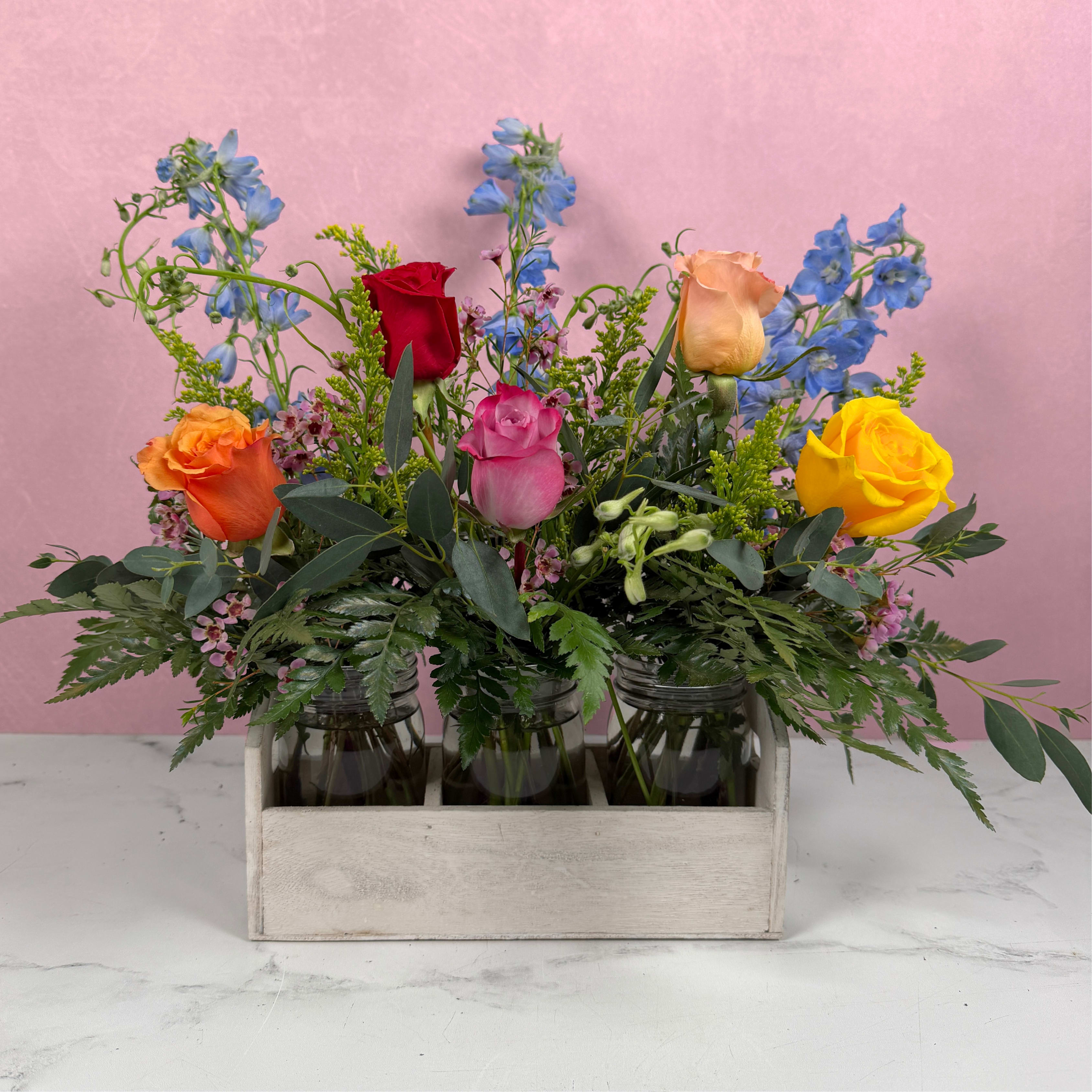 Celebrating You Mason Jar Trio - Bright, garden-fresh color greets the recipient at first glance: mixed roses pair with tall blue delphinium spikes for a lively, celebratory bouquet perfect as a birthday bouquet, thank-you gift, or everyday surprise. Presented in a rustic trio of mason jars nestled in a whitewashed wooden crate, this flower arrangement reads joyful and hand-tied-the kind of gift that makes someone feel truly noticed.  The design is full and layered, with fern-like foliage and eucalyptus adding soft texture around the base and small purple and pink filler blooms punctuating the color mix. The mason jar trio creates a casual, homey display that brings vibrant color to kitchens, office desks, or celebration tables.  Choose this arrangement for birthdays, congratulations, or a cheerful thank-you-it's the kind of bouquet that brightens a room and lifts spirits. This arrangement arrives ready to display for immediate enjoyment, and we recommend fresh water and a daily trim to keep blooms looking their best.
