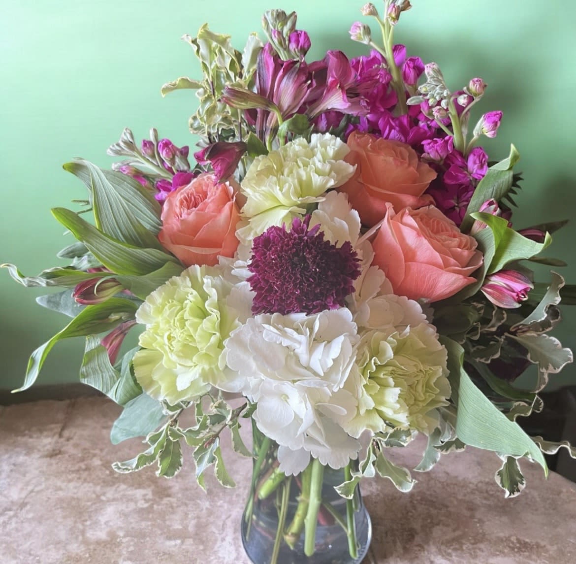 Blush Serenity  - A joyful bouquet of soft pinks and peaches that feels like a warm embrace - Blush Serenity is a flower arrangement perfect for birthdays, celebrations, or sending cheerful well-wishes. This birthday bouquet and celebration arrangement presents a fresh, uplifting palette of peach, pink, white, and soft green that will make the person you're thinking of feel genuinely seen and celebrated.  At the center, a dense cluster of pale green and white carnations and hydrangea creates a lush, textured core, punctuated by a single dark burgundy bloom for striking contrast. Peach roses form a gentle ring around the middle while bright pink alstroemeria and upright stock add height and lively movement. All stems are gathered in a simple clear glass vase that showcases the straight stems and gives the design a clean, classic silhouette.  Ideal for friends, family, or coworkers who deserve a cheerful surprise, this arrangement reads as both elegant and approachable - the kind of gift that brightens a room and lifts a mood. Vase dimensions and care: keep water fresh, trim stems every few days, and place away from direct heat or drafts to extend blooms; standard vase sizing fits a medium tabletop display.